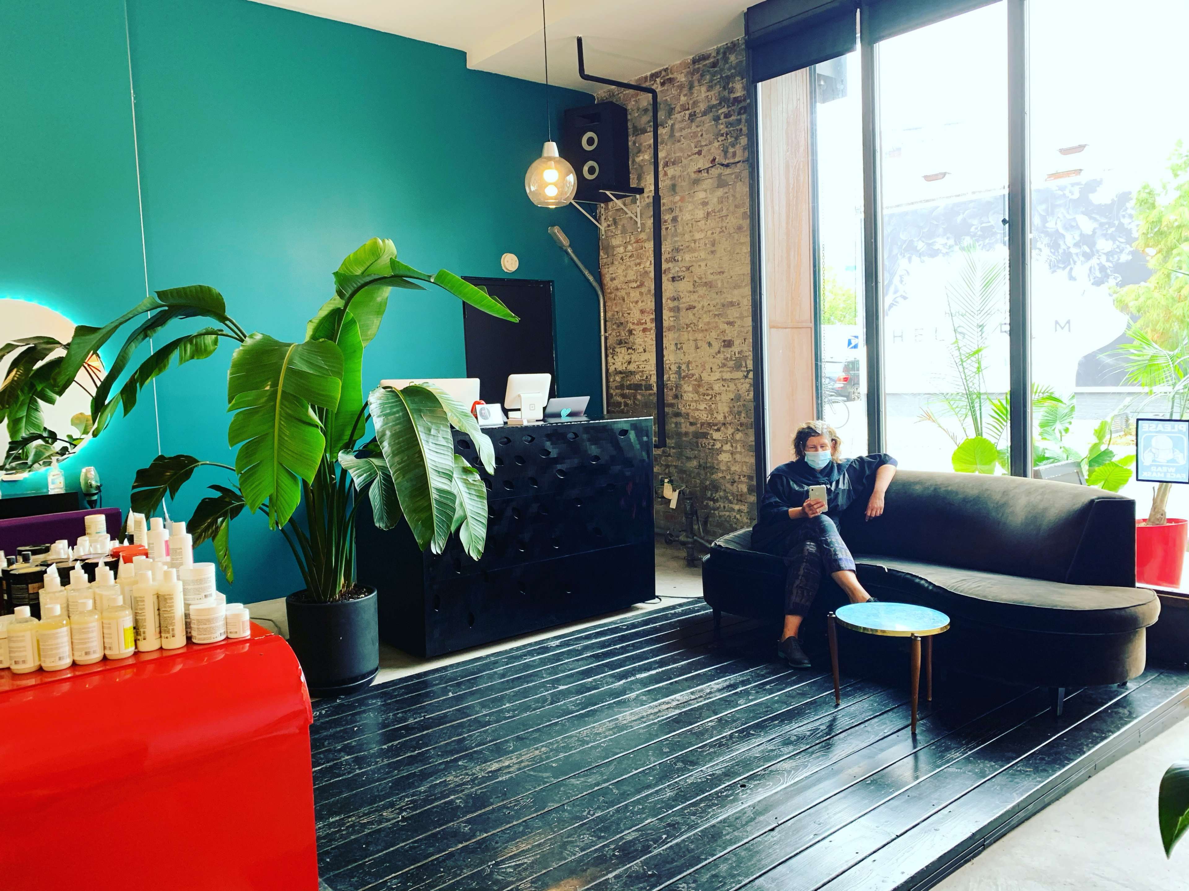 A modern salon features a sleek black counter, a comfortable black sofa, and a large window with natural light illuminating the space, surrounded by green plants and beauty products displayed on a table.