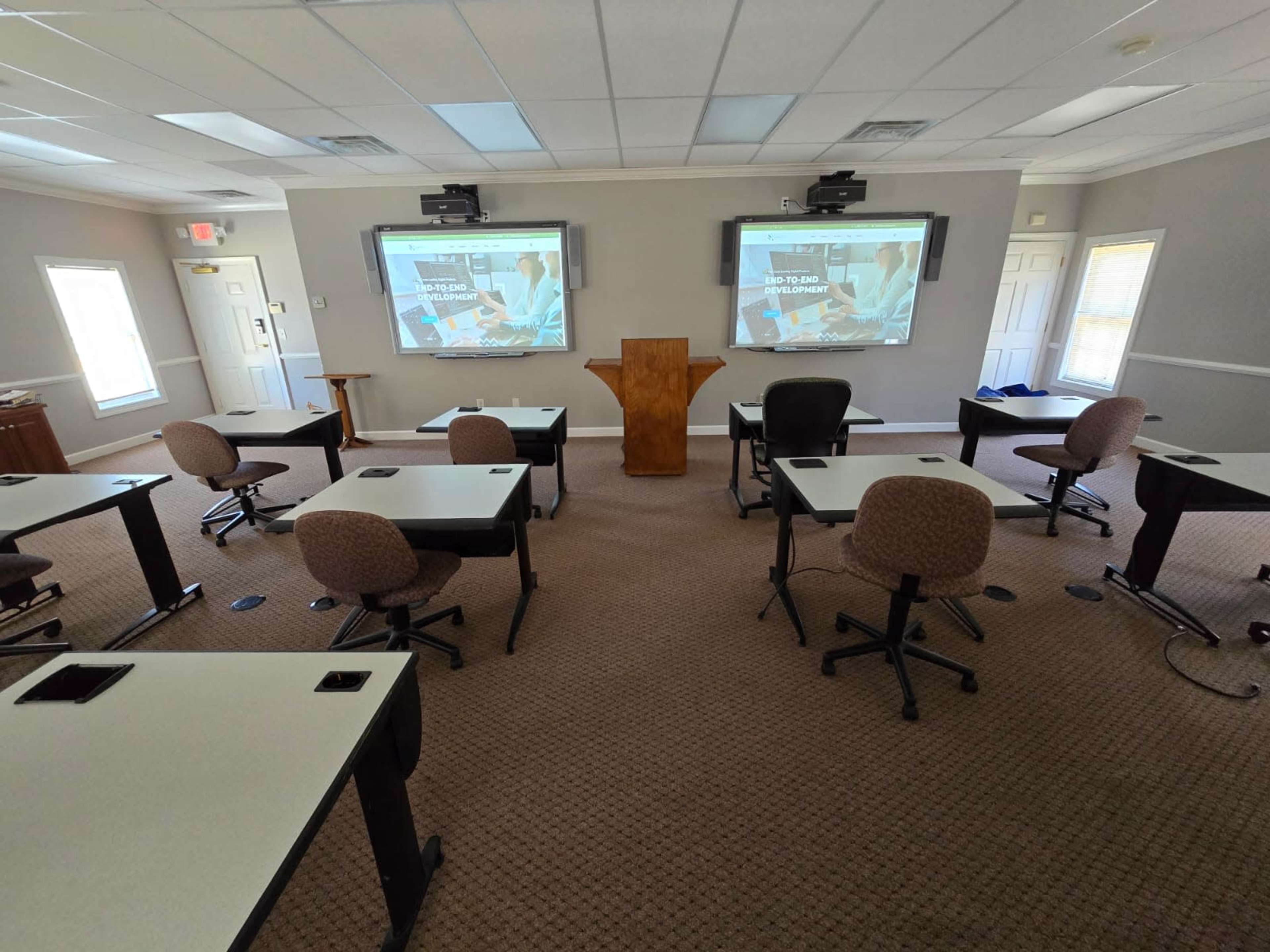 A classroom is set up with white tables and rolling chairs facing two large screens displaying presentation slides.