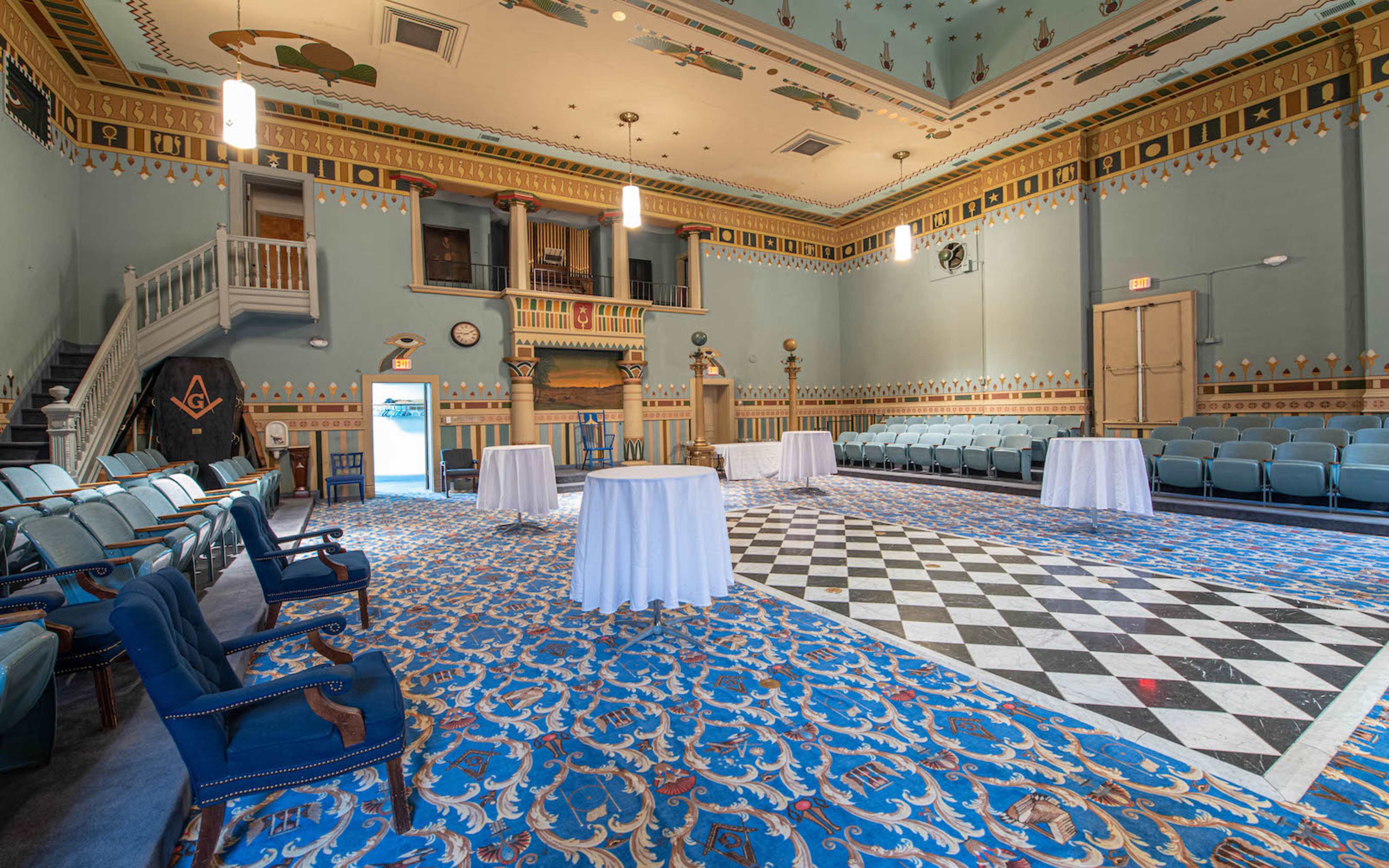 A spacious, well-decorated room with a blue tiled floor featuring ornate designs, several round tables covered in white tablecloths, and a checkered area in the center, framed by green chairs and a staircase.