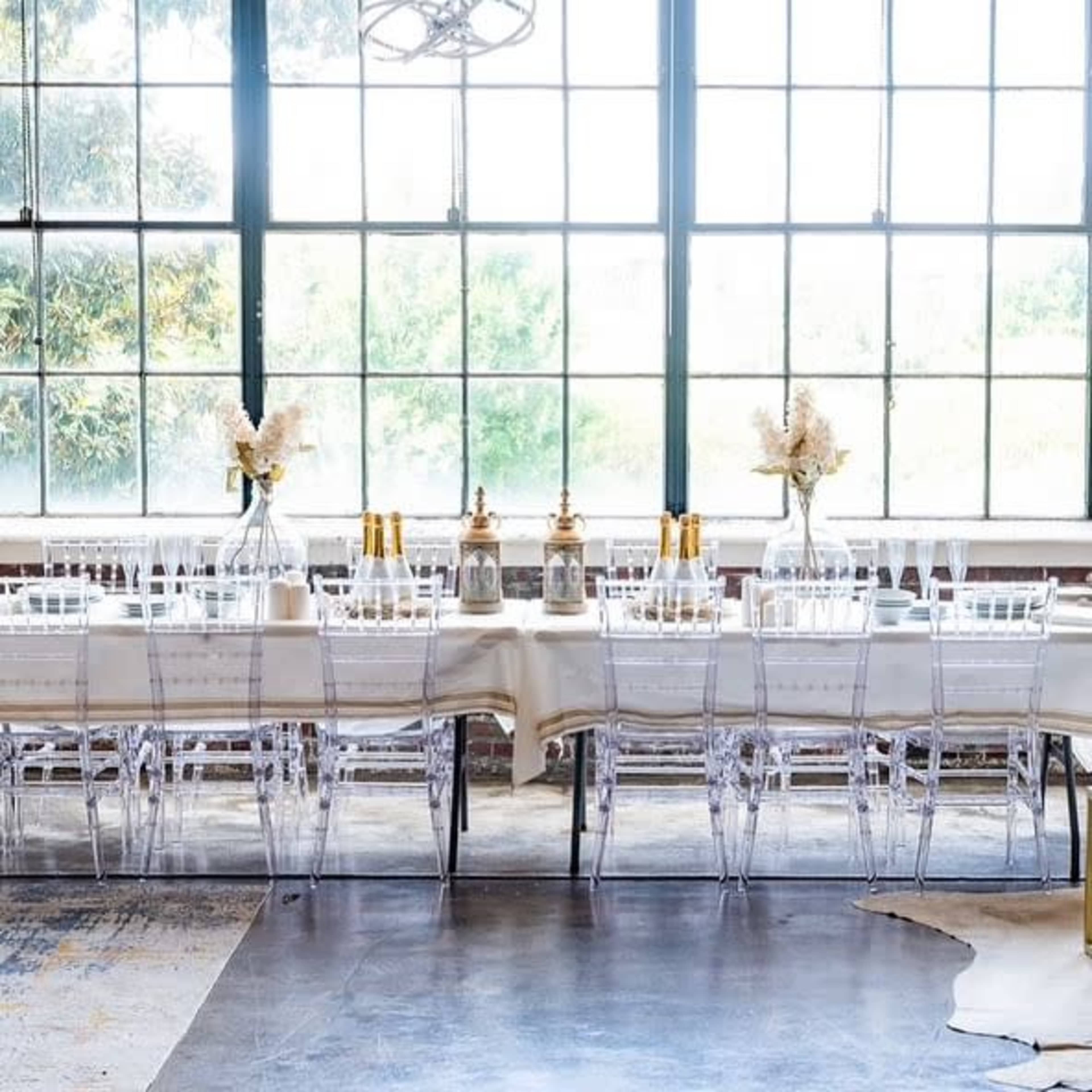 A long dining table is set with clear chairs, glassware, and decorative vases, positioned near large windows that let in natural light.