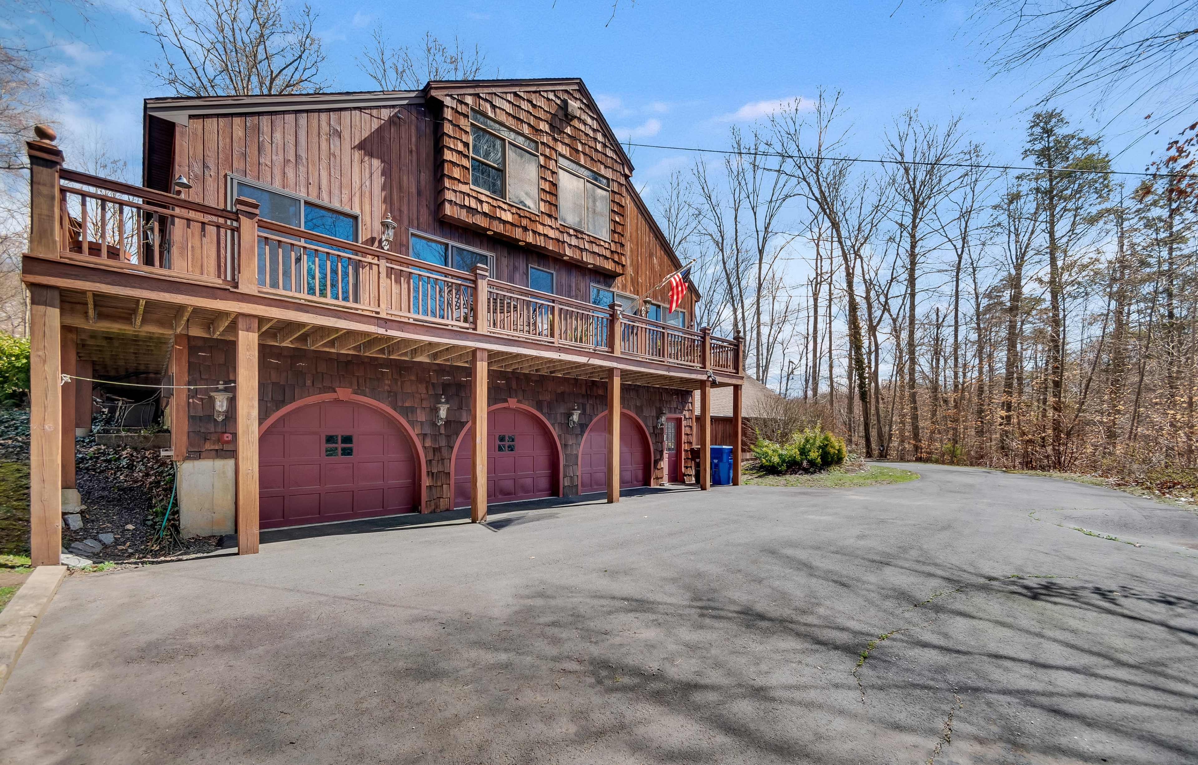 A large wooden house with a sloped roof, featuring three garage doors at the ground level and a balcony above, surrounded by a forested area in early spring.