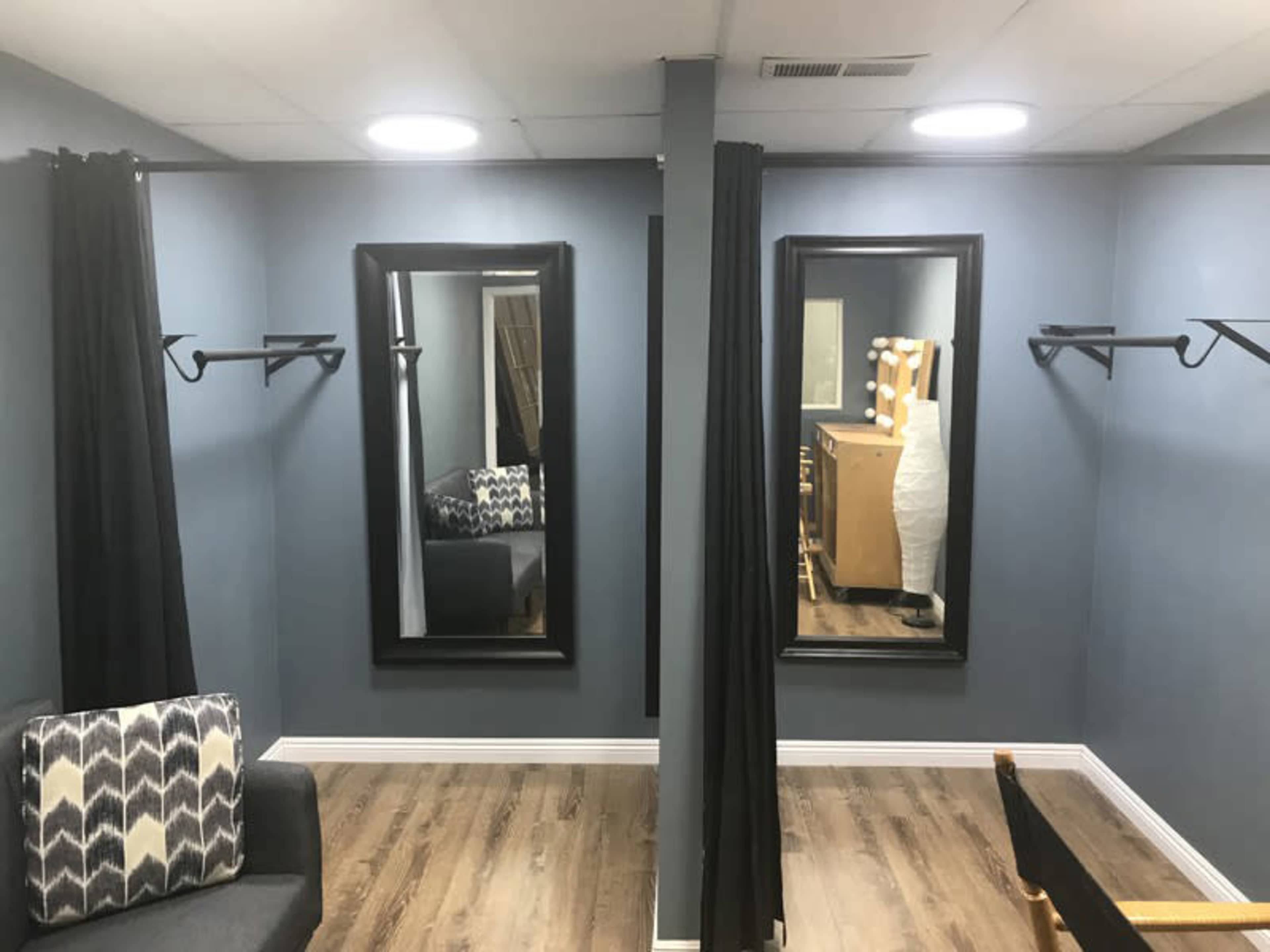 A dressing room with two large mirrors, dark curtains, a gray couch, and a light-colored wooden dresser.