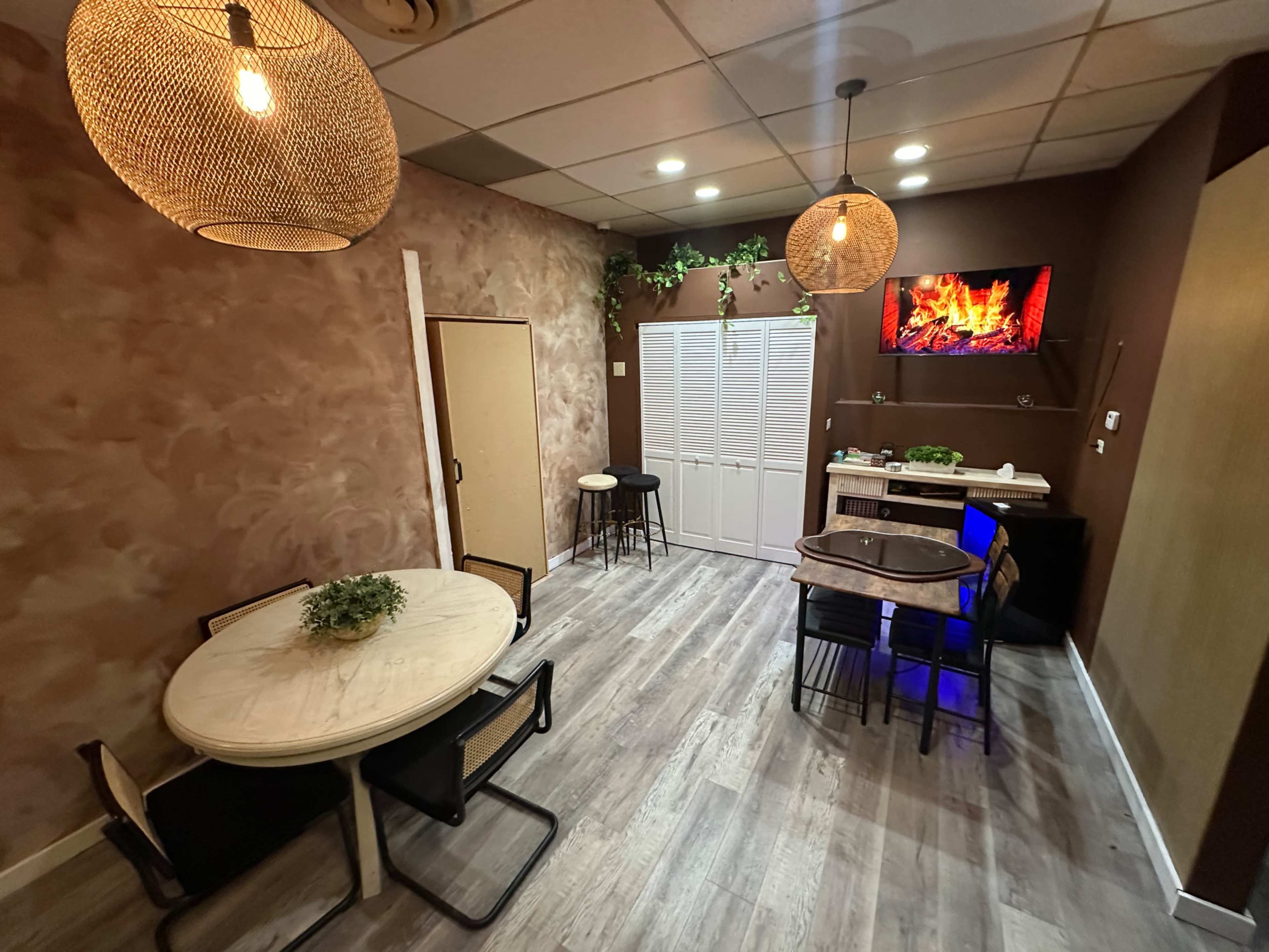 The image shows a cozy interior room with a table, chairs, and decorative lighting, featuring a wall-mounted screen displaying a fireplace effect.