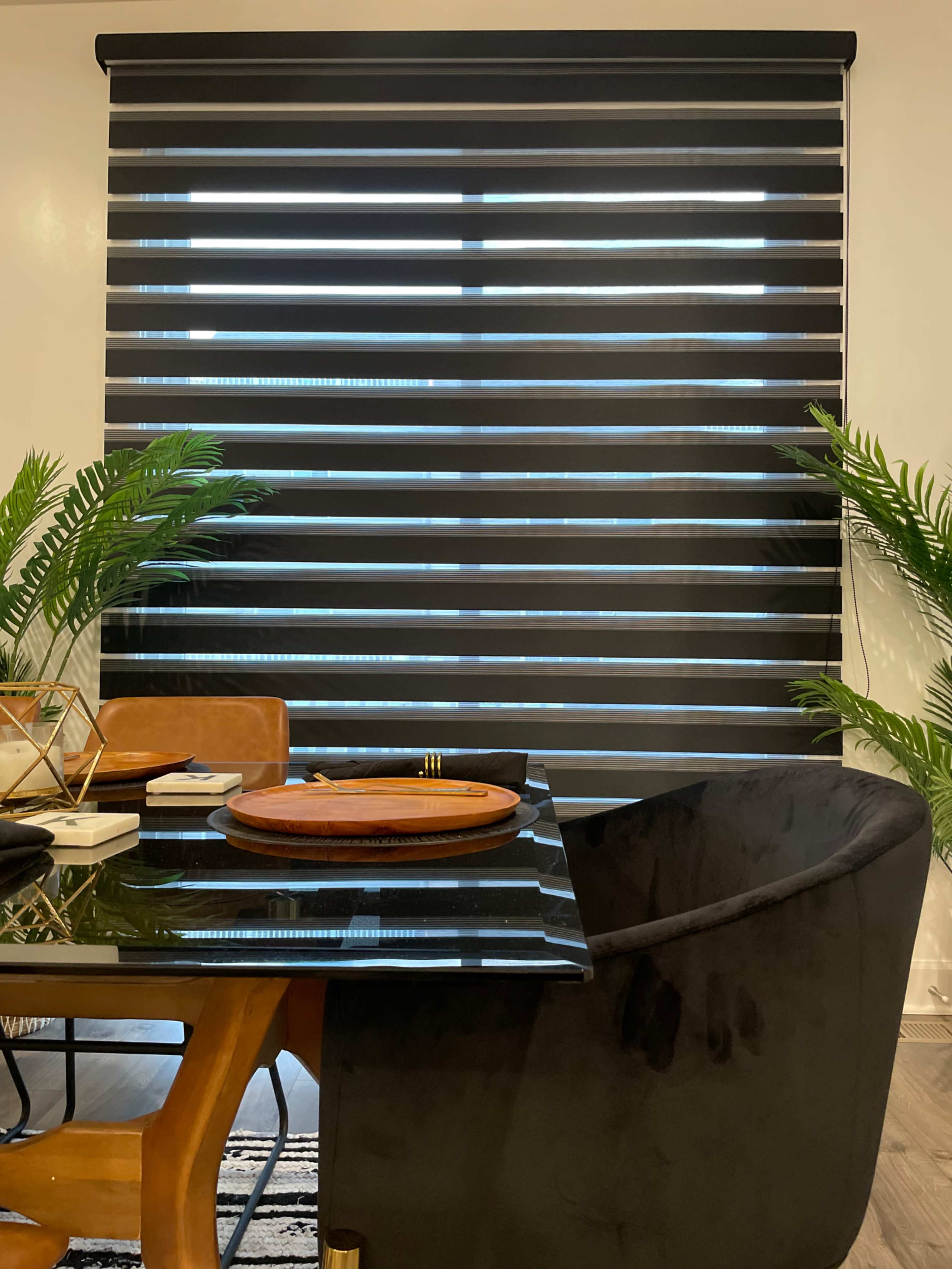 A modern dining area features a black and white striped window treatment, a glossy black table, and two green plants in the foreground.