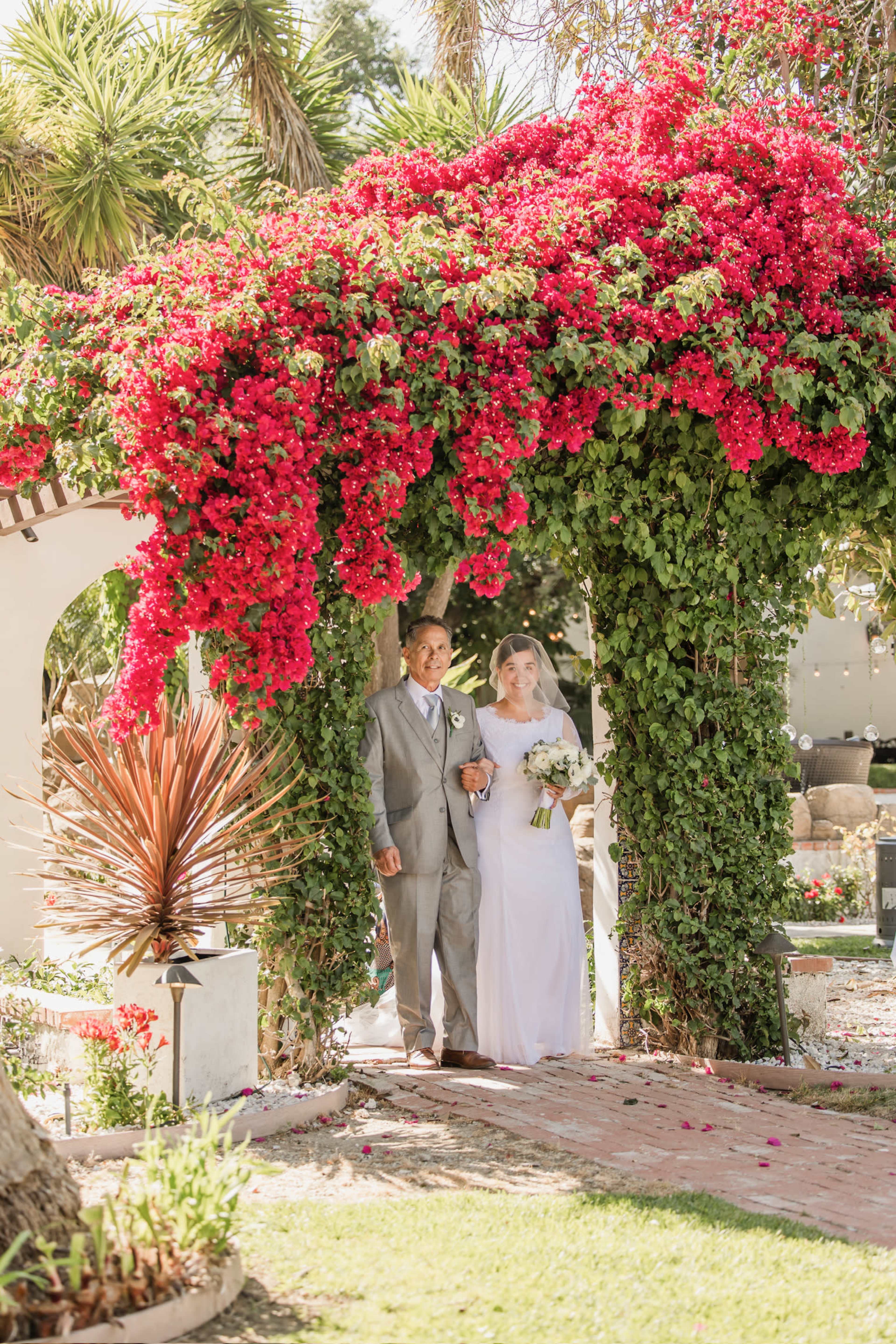 A man and woman stand under a floral arch adorned with vibrant red bougainvillea at a wedding venue.