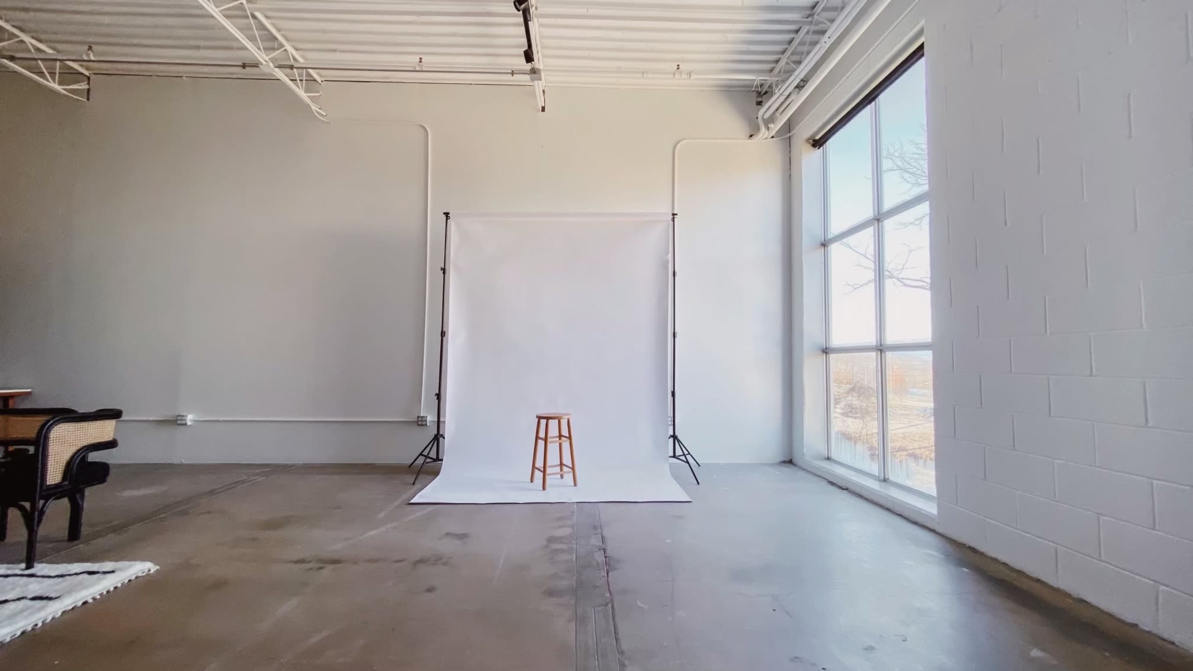 A spacious indoor studio with a plain white backdrop, a wooden stool positioned in front of it, and large windows allowing natural light to enter.