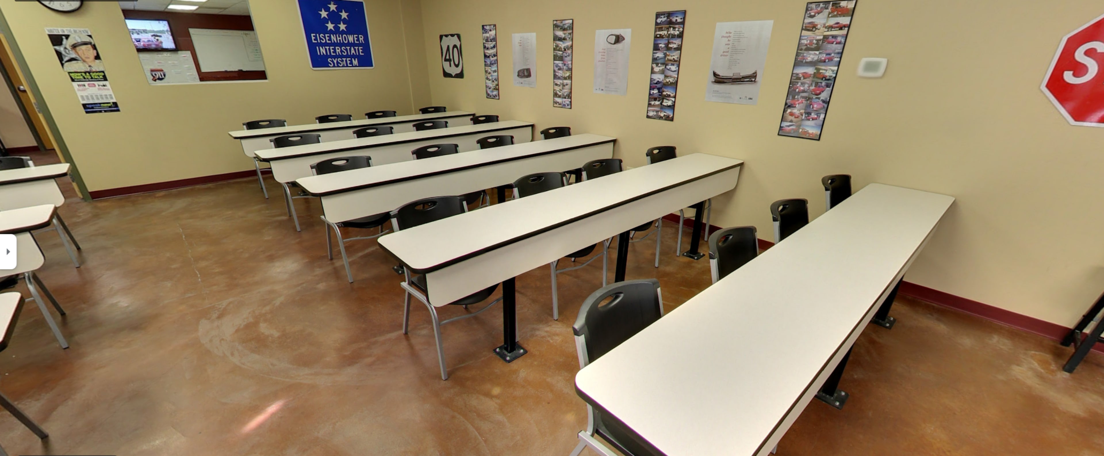 The image shows a classroom arranged with several rows of long tables and black chairs, featuring educational posters on the walls.