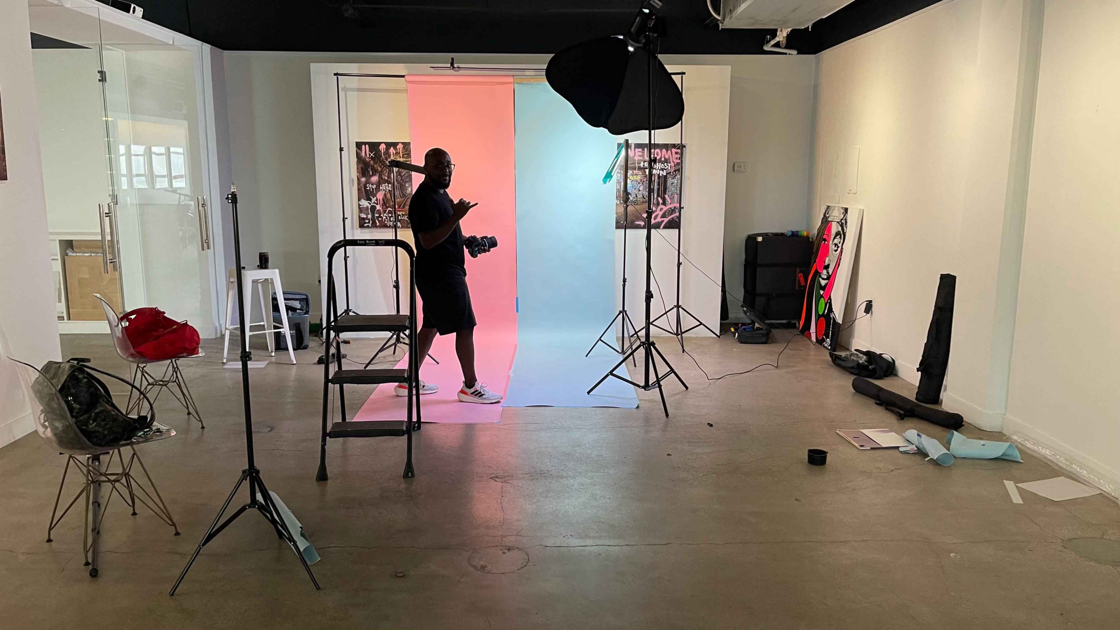 A person stands on a ladder in a studio setting with a pink and blue backdrop, surrounded by photography equipment and various props.