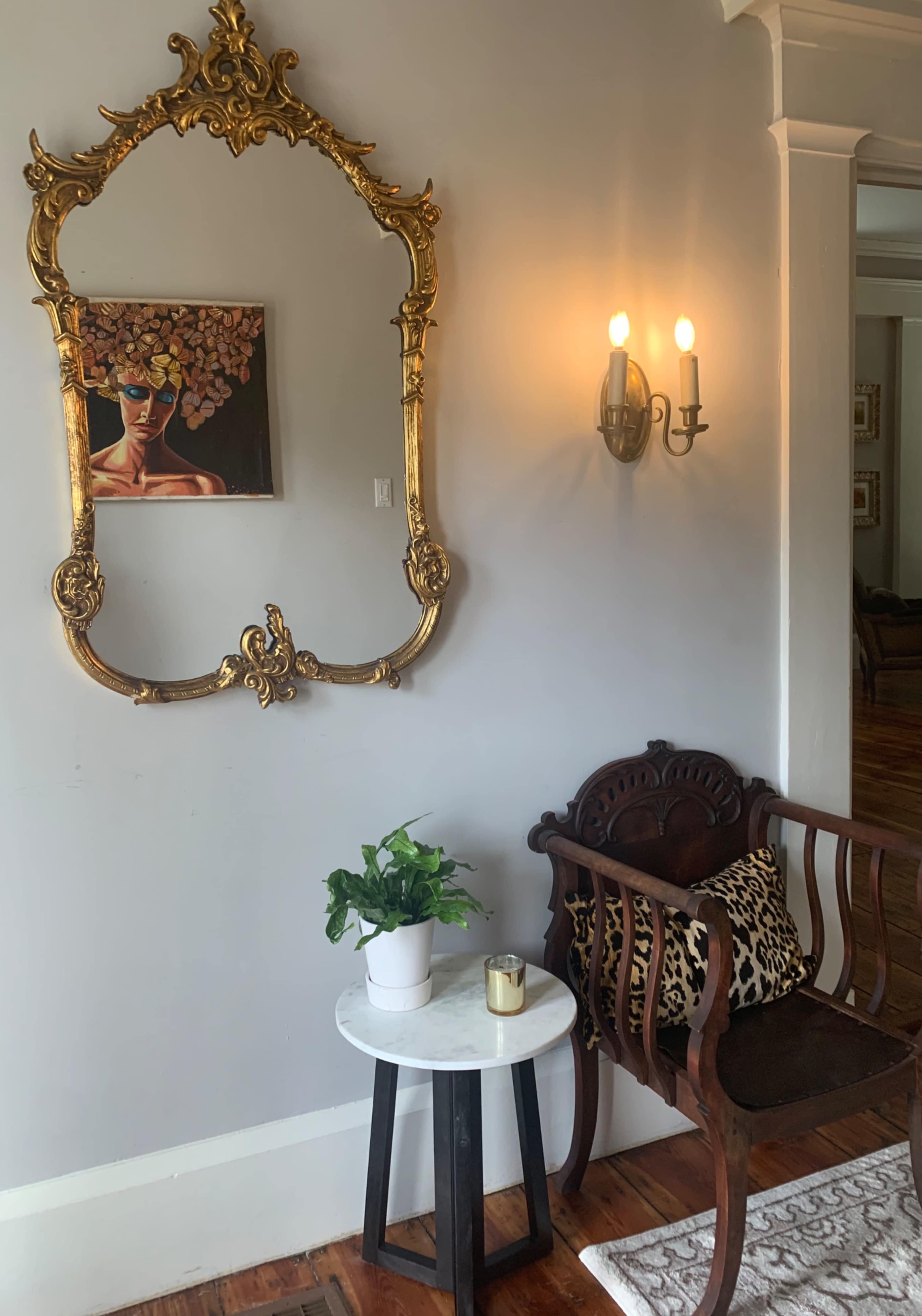 The scene features a wall with a large ornate mirror and a painting, alongside a wooden bench with a patterned cushion, a small round table, a potted plant, and a candle.