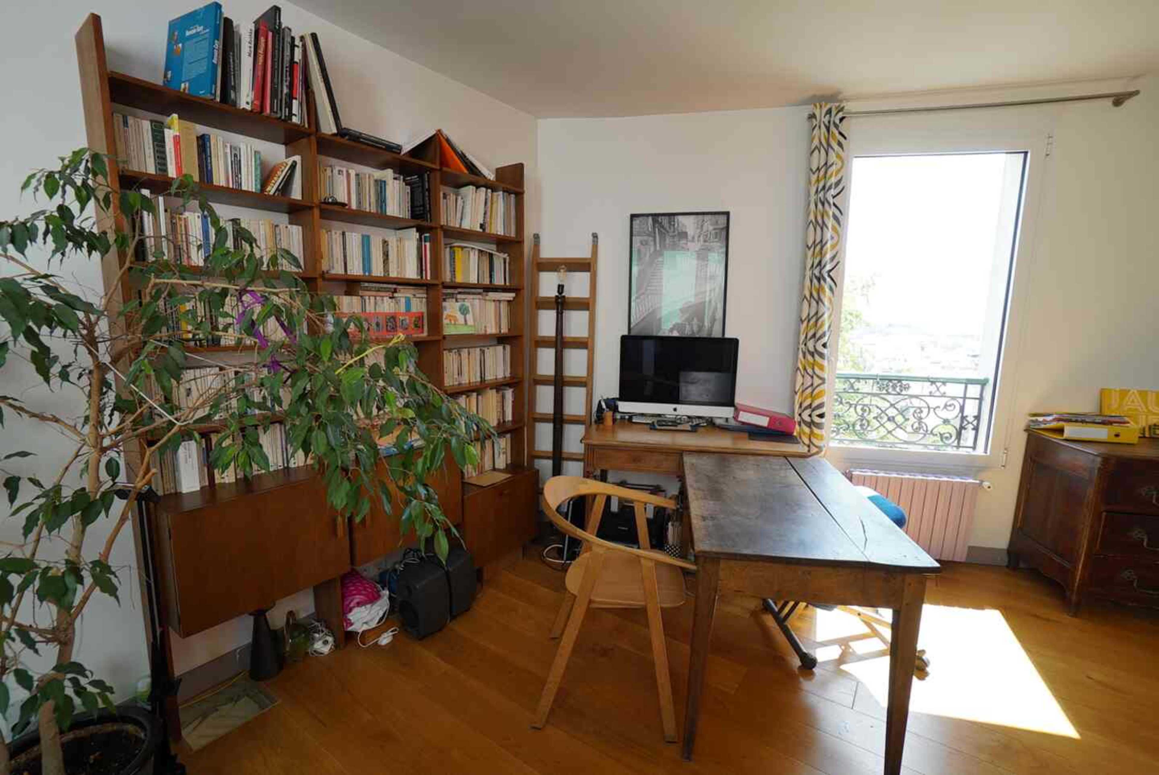 The image shows a cozy room with a wooden desk, a computer, a bookshelf filled with books, and a potted plant near a window.