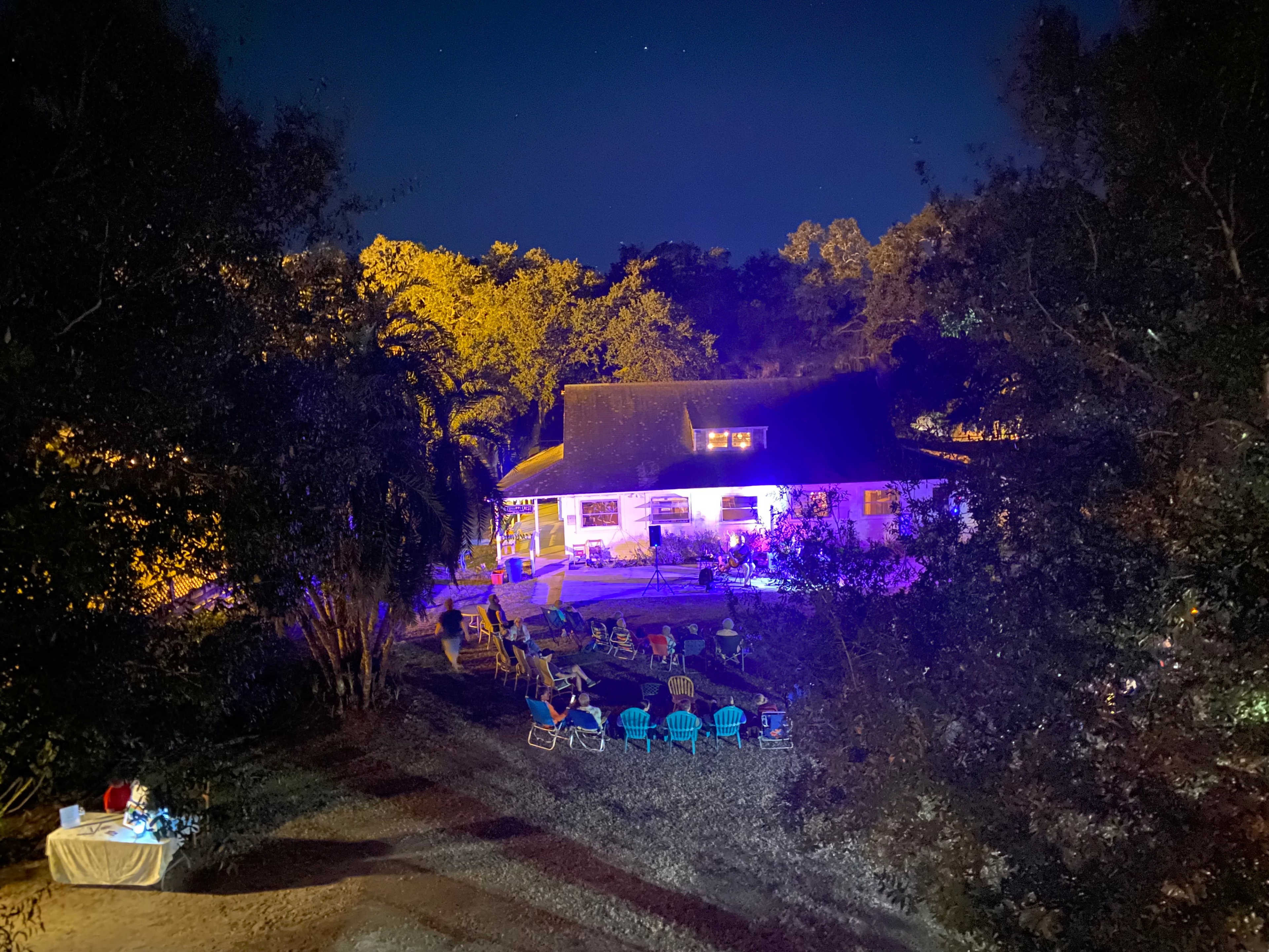 A house is illuminated by colorful lights at night, surrounded by chairs arranged on a grassy area amidst trees.