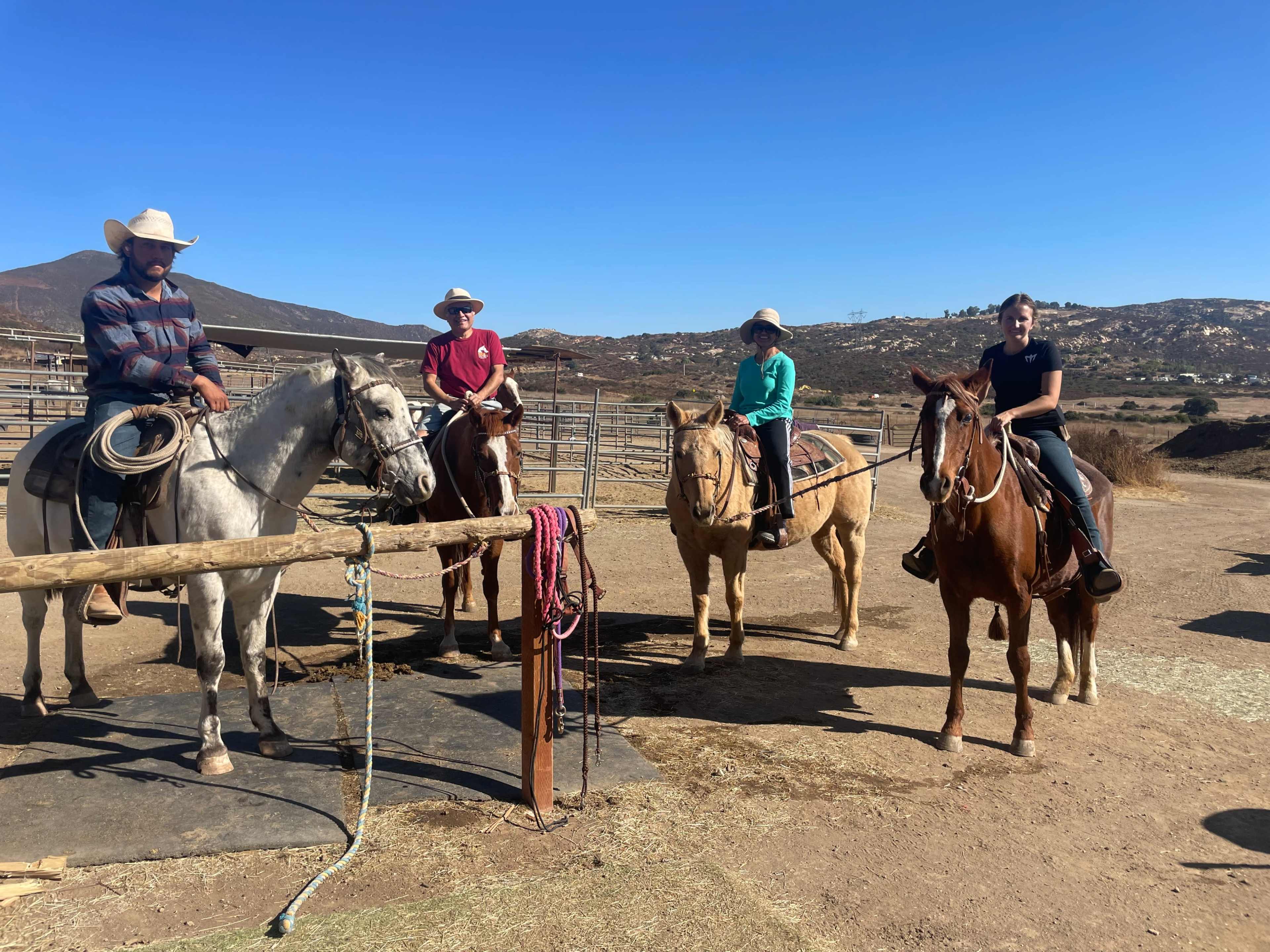 Four individuals are mounted on horseback in a corral under a clear blue sky.