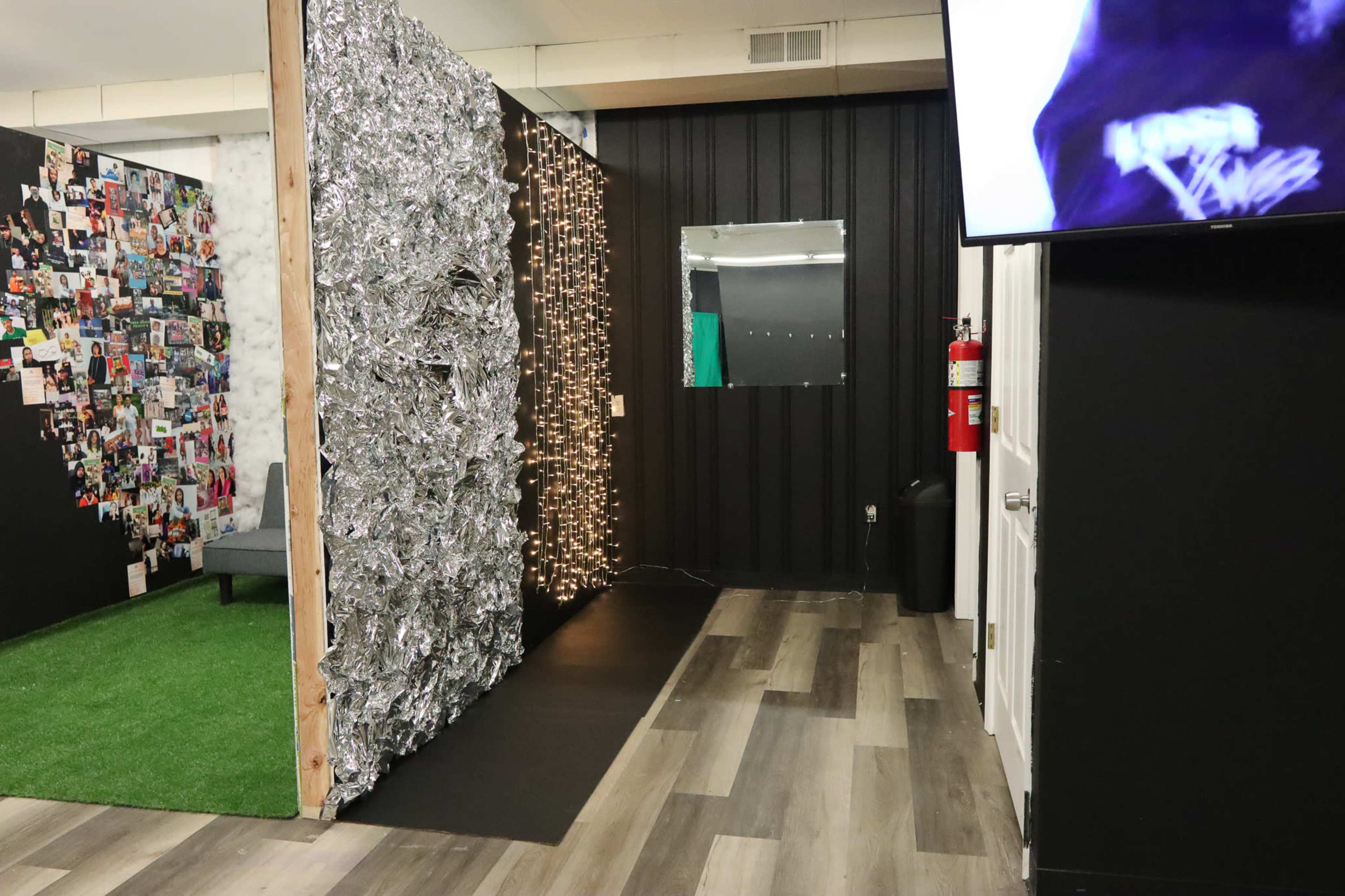 An interior space with a textured wall made of shiny material, a large collage of photos on the left, and a mirror on the right, alongside a TV mounted on the wall.