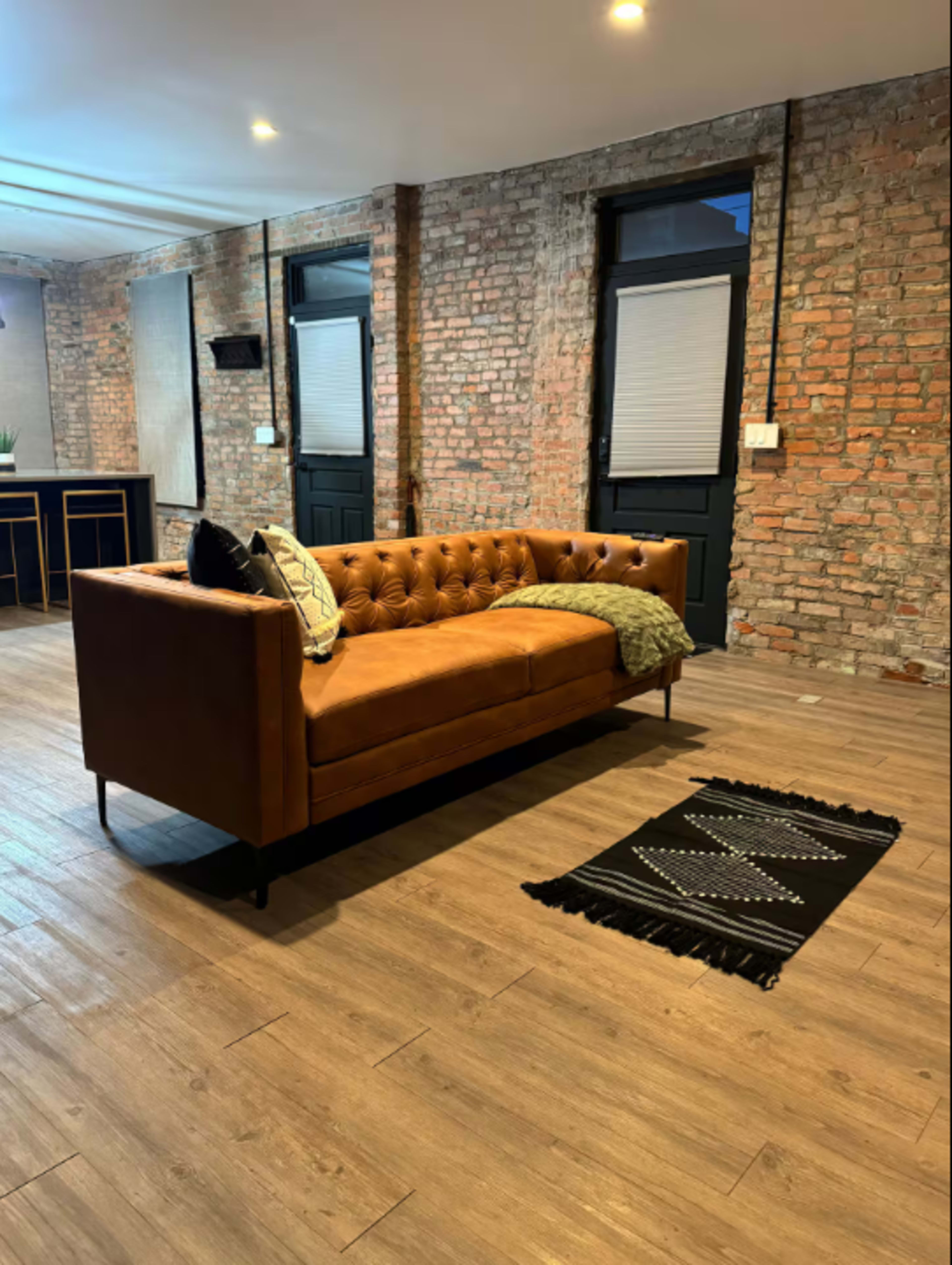 The image shows a brown tufted leather sofa positioned on a wooden floor in a room with exposed brick walls and two doors in the background.