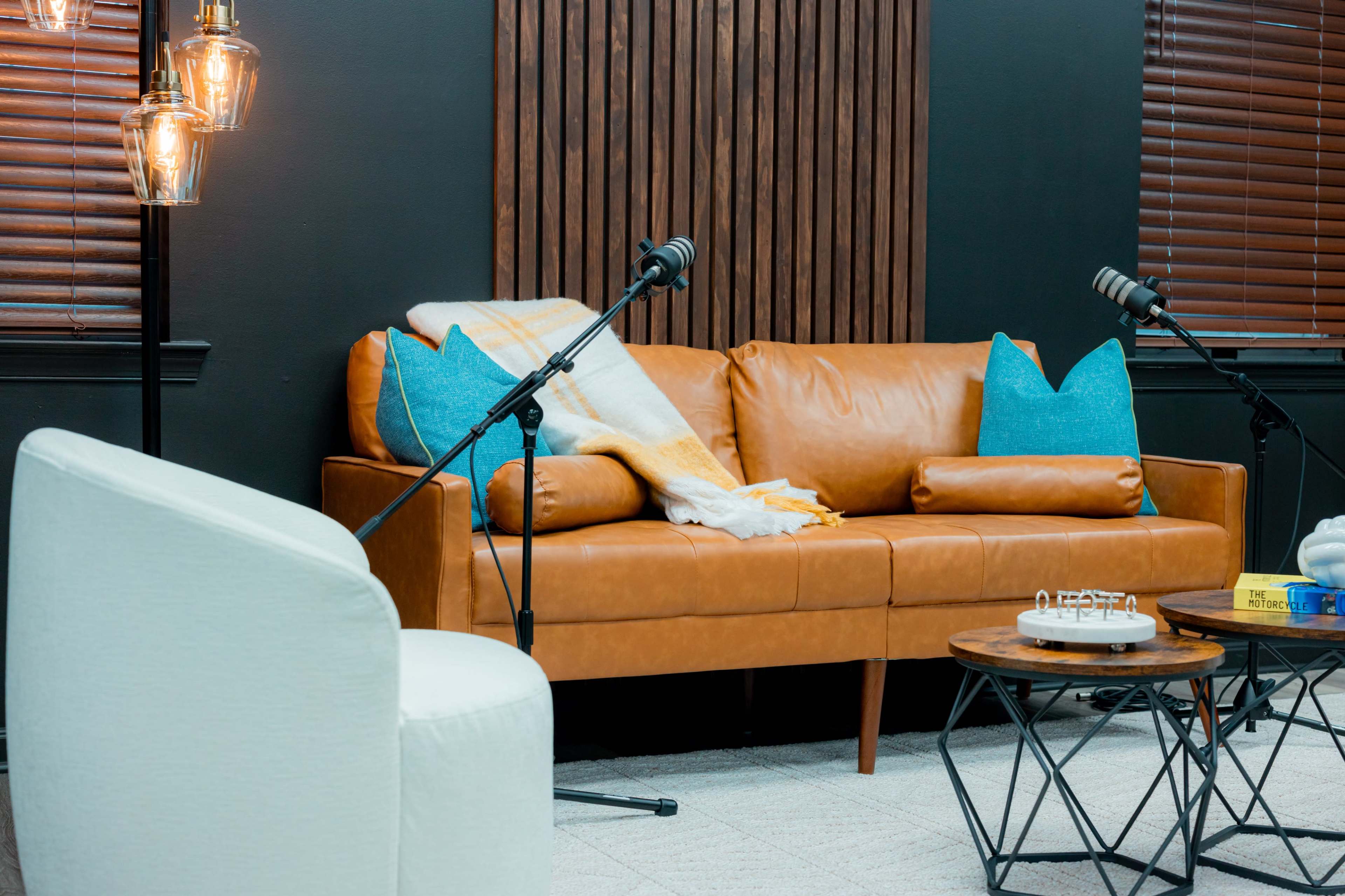 The image shows a cozy seating arrangement with a brown leather sofa adorned with turquoise pillows and a white armchair, accompanied by microphones on stands and a wooden coffee table.