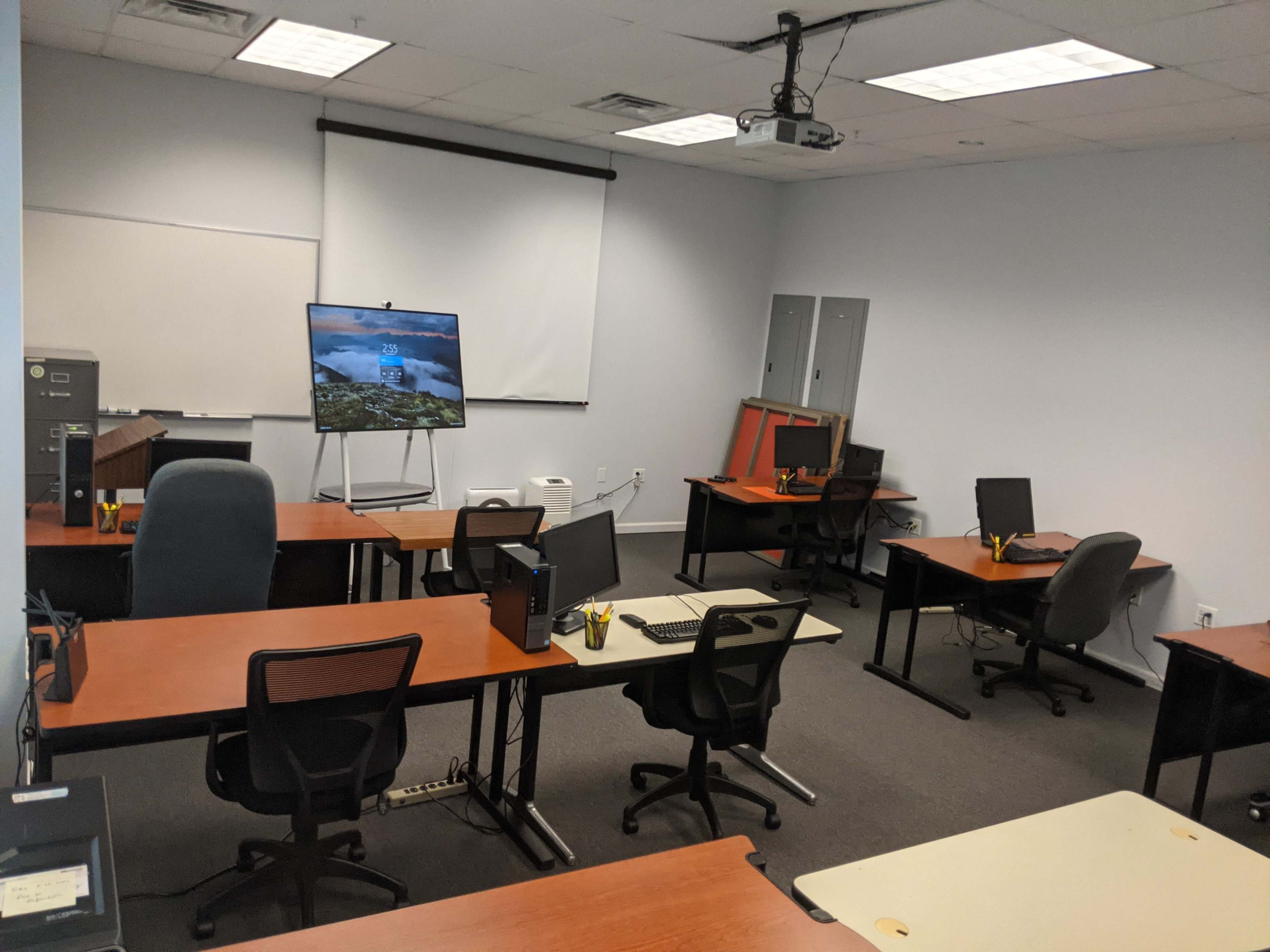 A classroom with several empty desks and chairs, a large screen on a stand, and a projector setup.