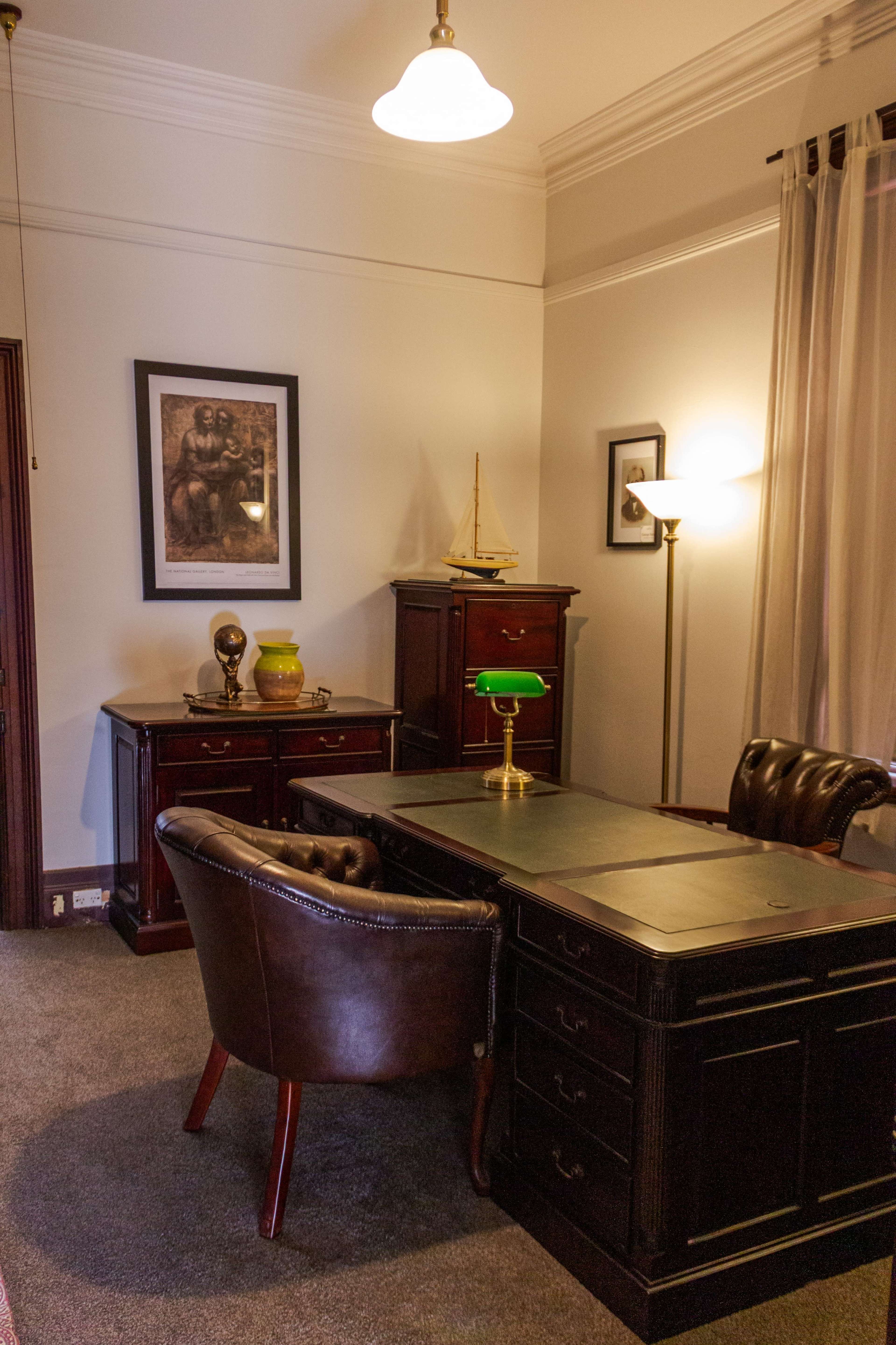 The image shows a classic office space featuring a dark wood desk, a leather chair, and decorative items, including a framed picture and a small boat model.