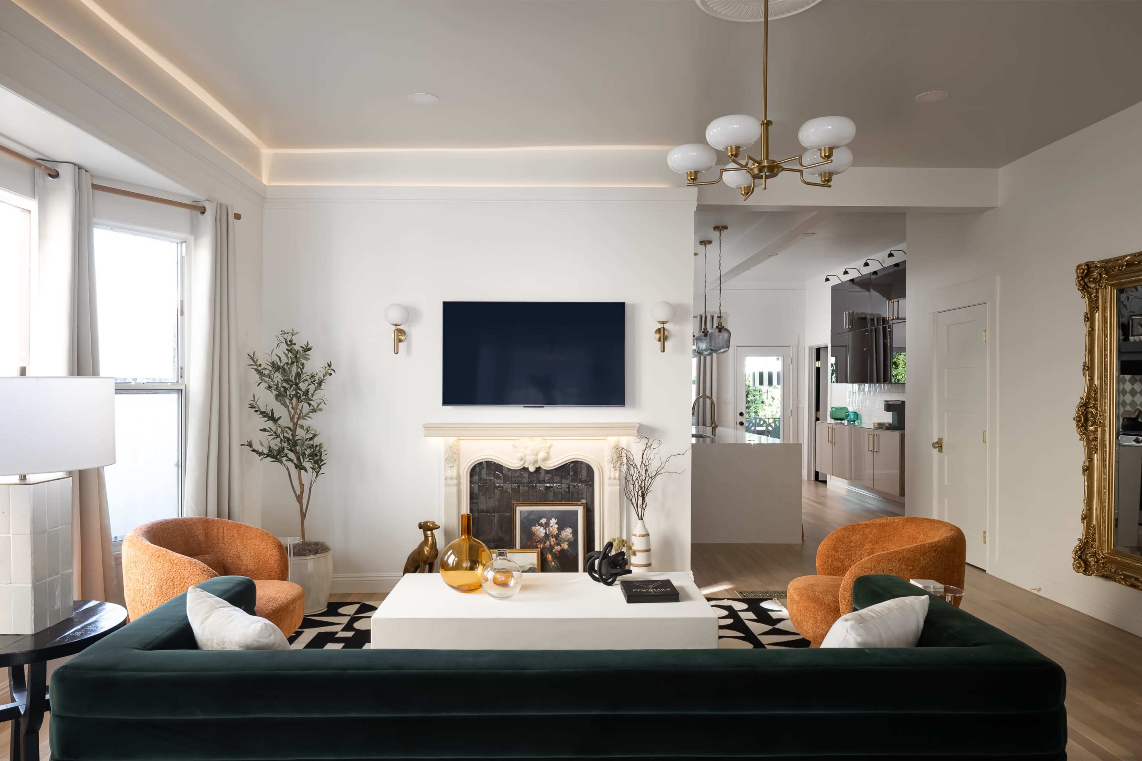 A modern living room features a green sofa, two orange armchairs, a white coffee table, and a wall-mounted plasma TV above a fireplace.