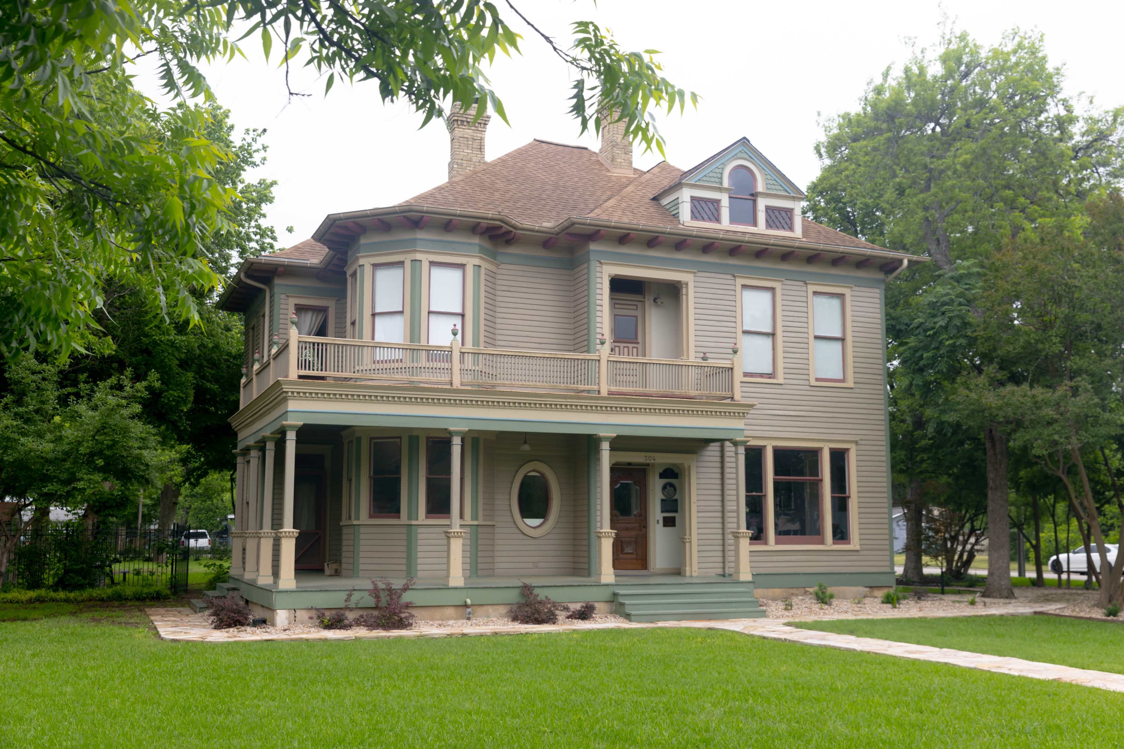 A large, two-story Victorian-style house with a wraparound porch and multiple windows sits on a grassy lawn surrounded by trees.