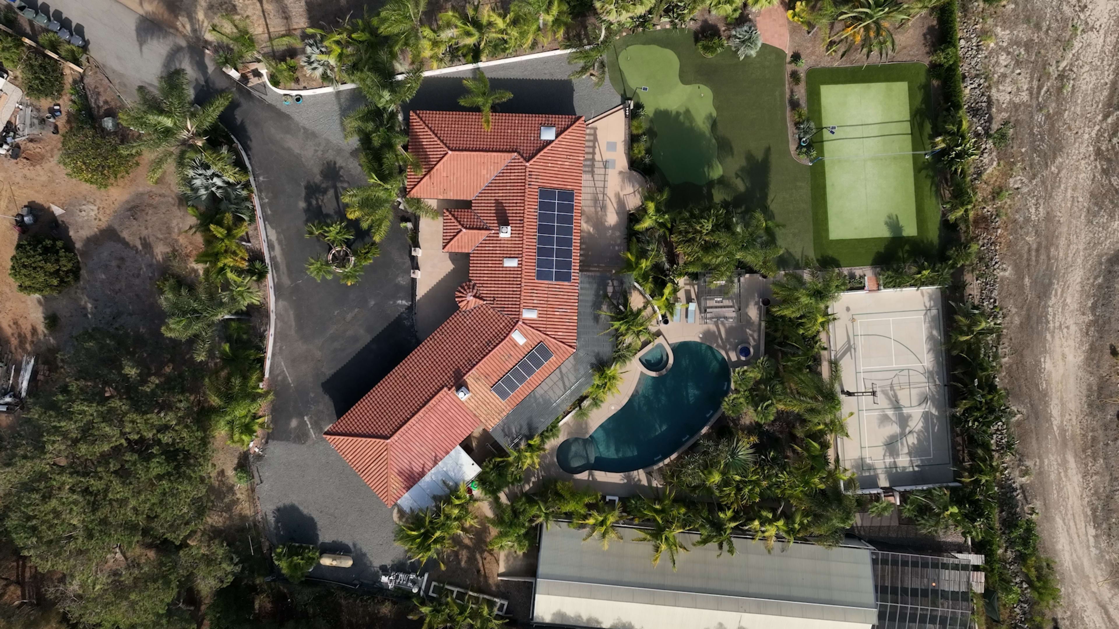 The image shows an aerial view of a residential property featuring a house with a red tiled roof, a swimming pool, a green lawn, and a sports court.