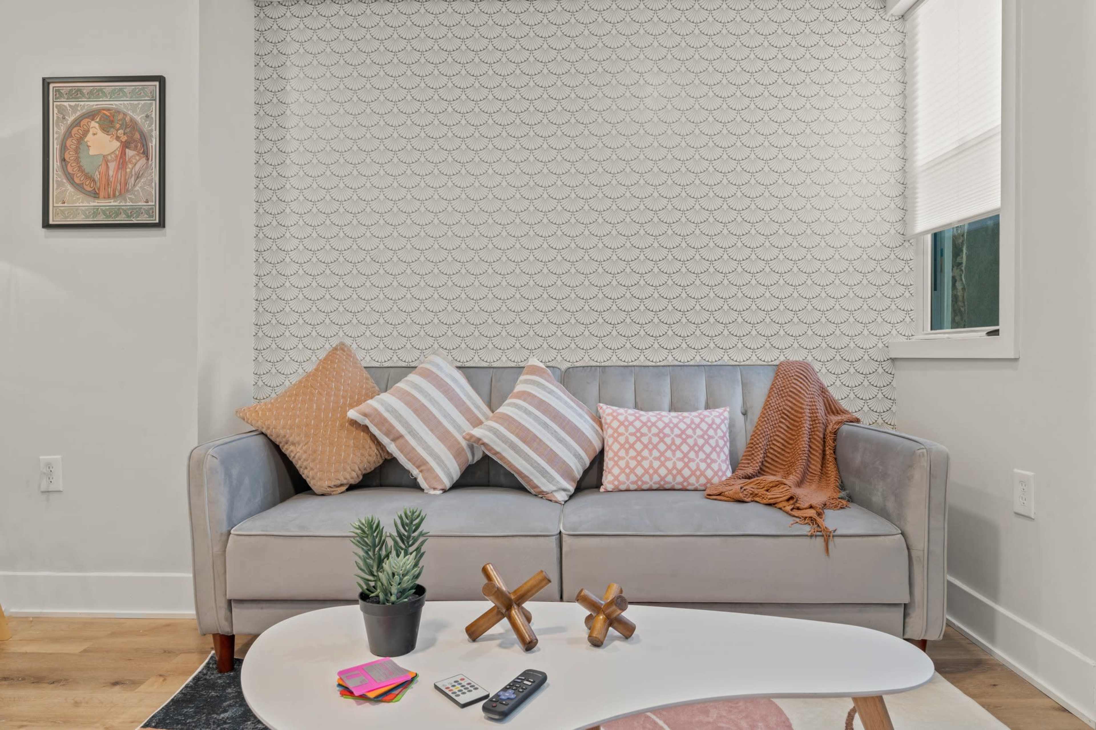 The image shows a modern living room with a gray sofa adorned with various colored pillows, a small table with decorative items, and a patterned wall in the background.
