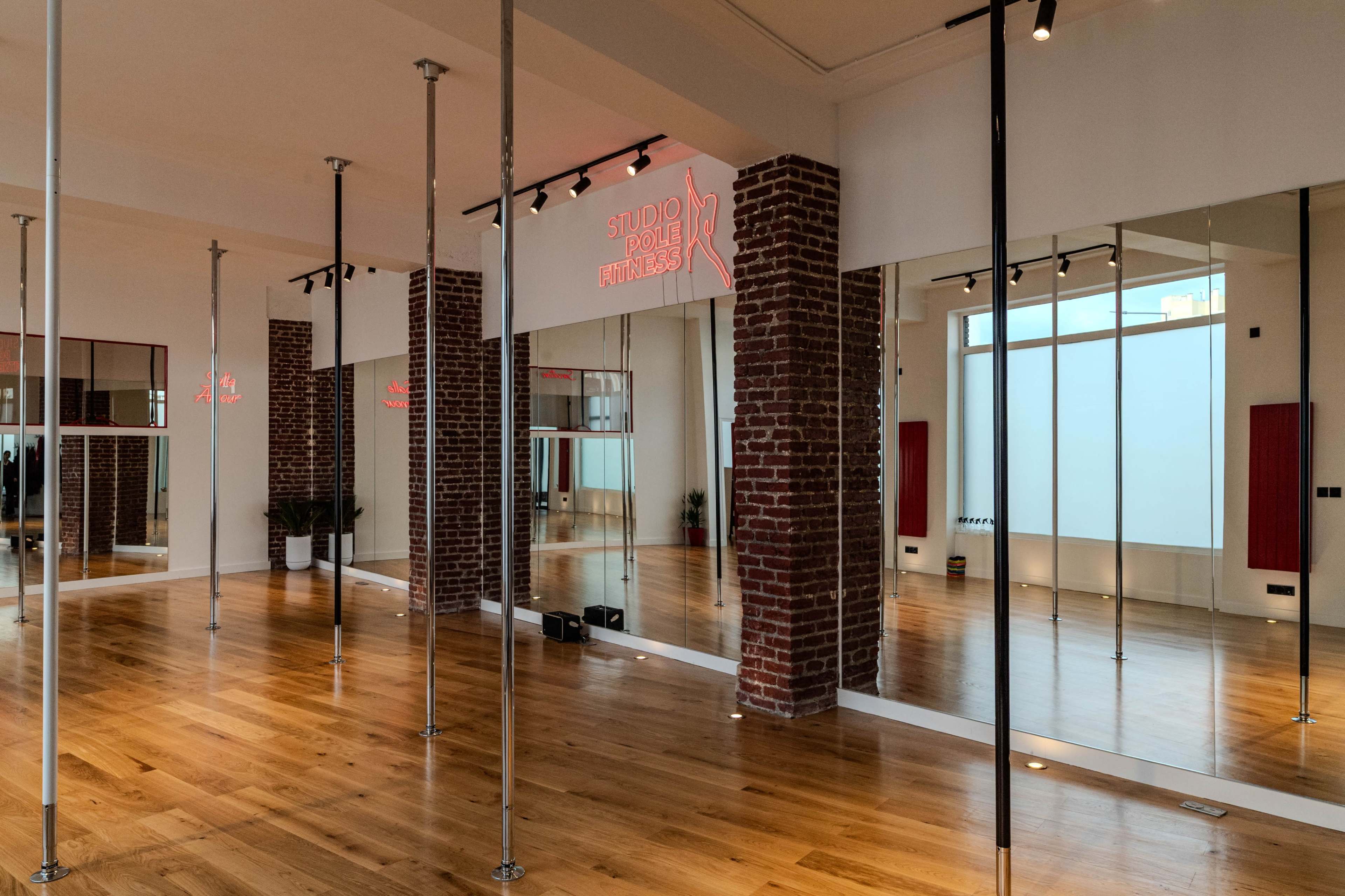 The image shows a fitness studio with wooden flooring, exposed brick walls, and mirrored panels, featuring poles for pole dancing.