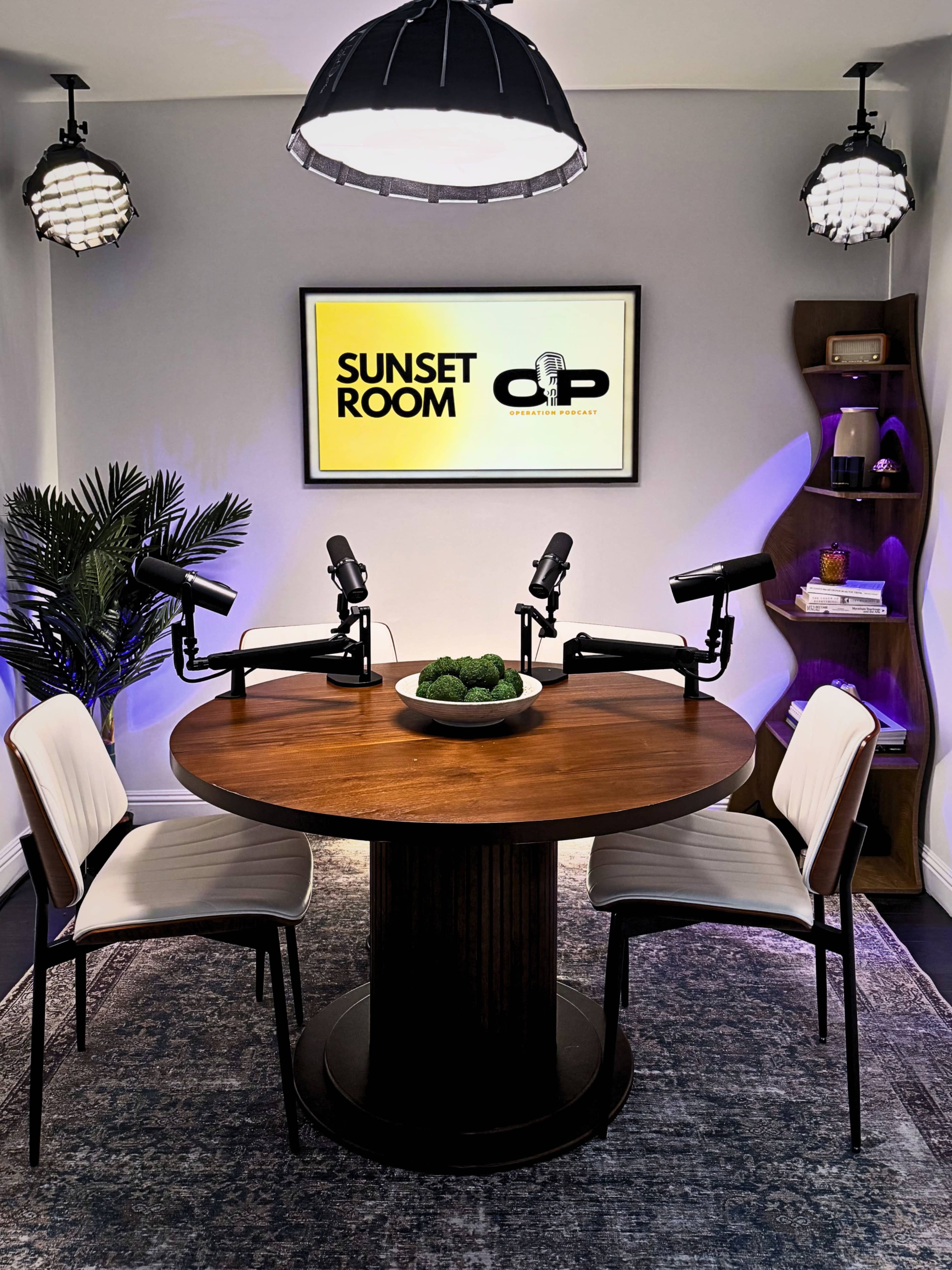 A modern podcast studio is set with a round wooden table, two empty chairs, and microphones arranged at the center, with a sign labeled "SUNSET ROOM" on the wall behind.