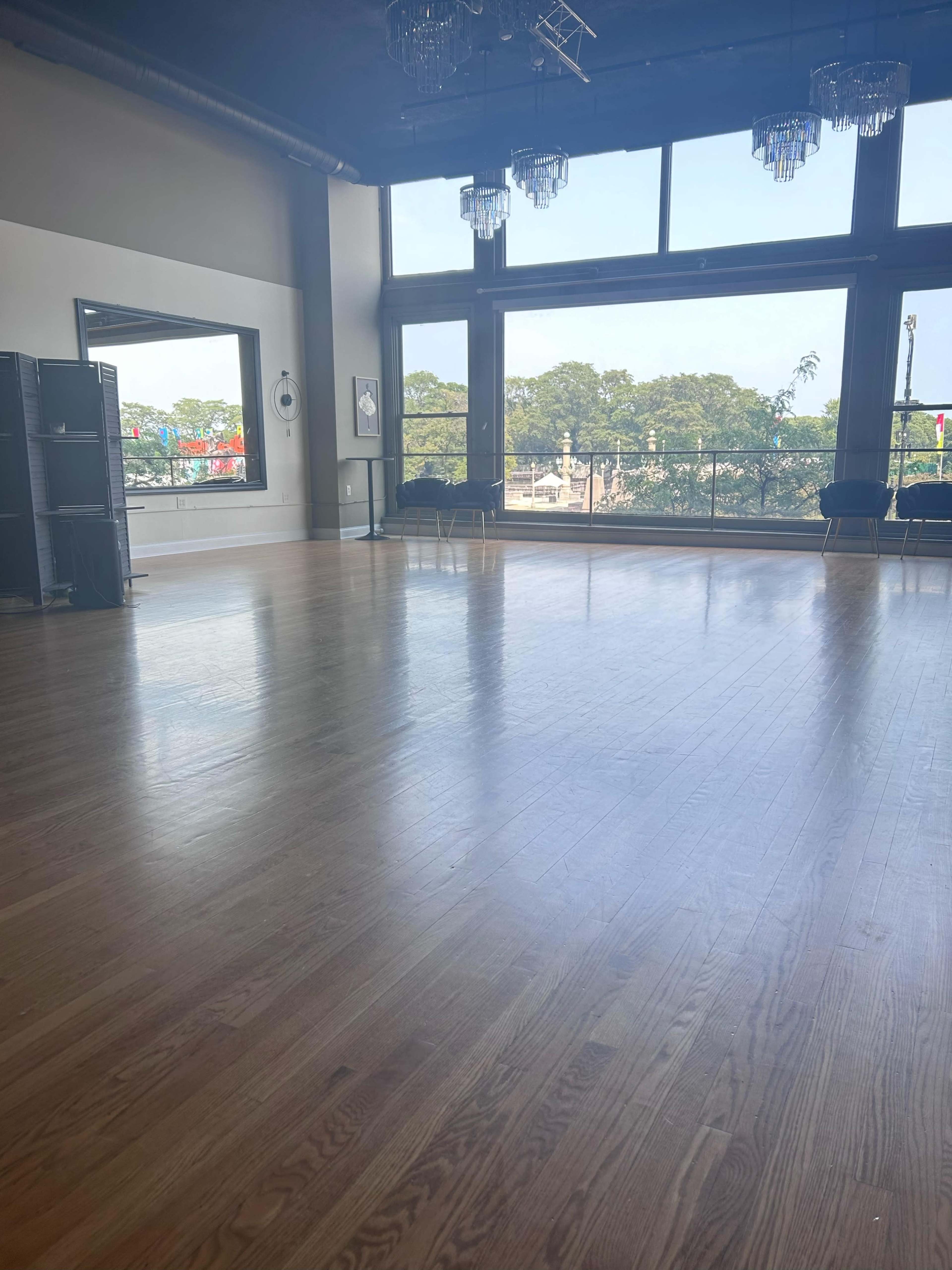 A spacious, empty dance studio with wooden flooring and large windows overlooking a tree-lined street.