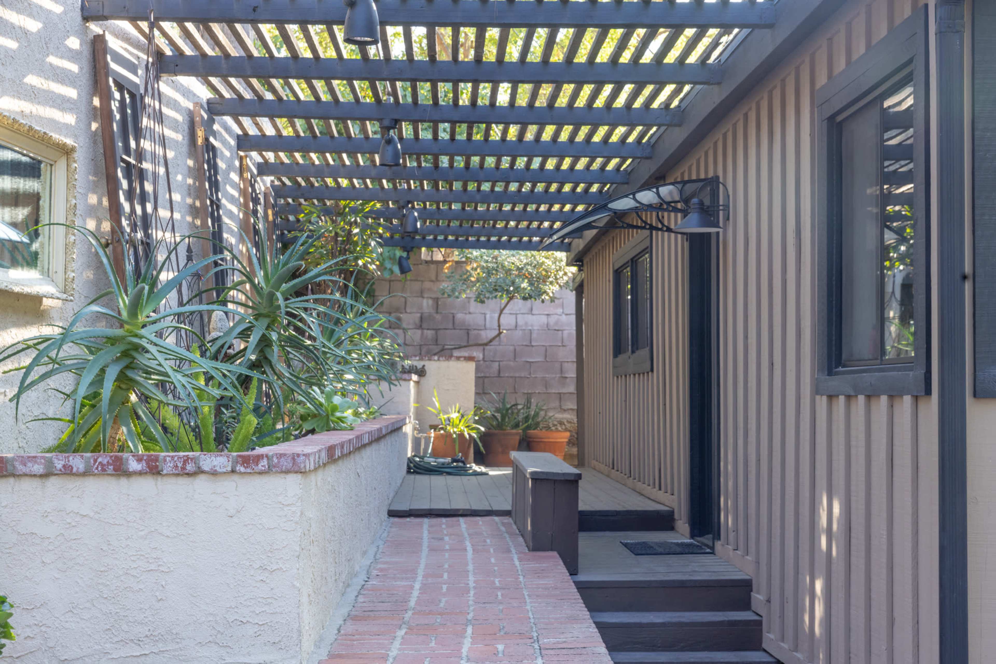 A narrow walkway runs alongside a house, featuring a pergola overhead and bordered by potted plants.