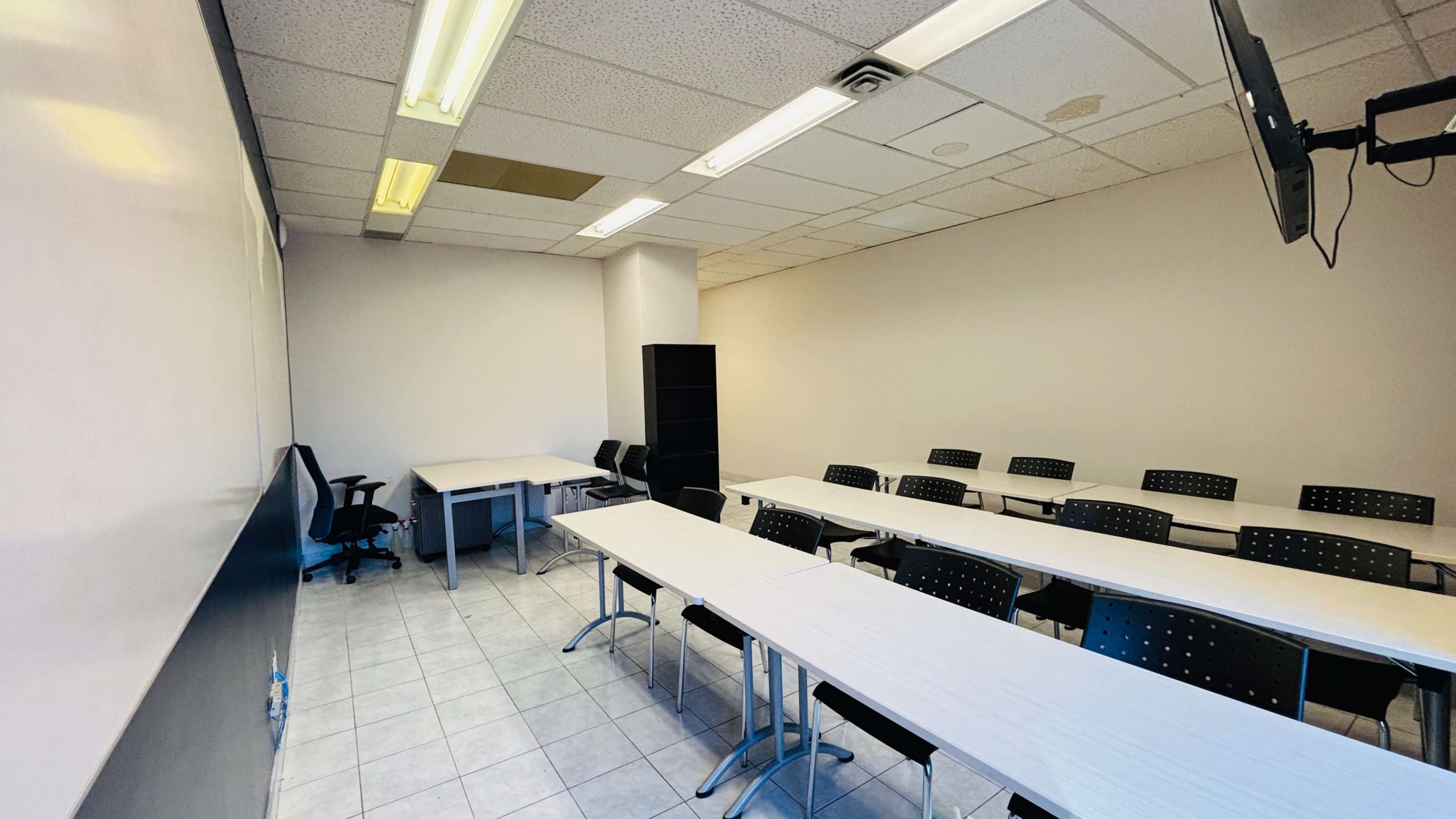 The image shows a vacant classroom with rows of tables and chairs, a black bookshelf, and a wall-mounted screen.
