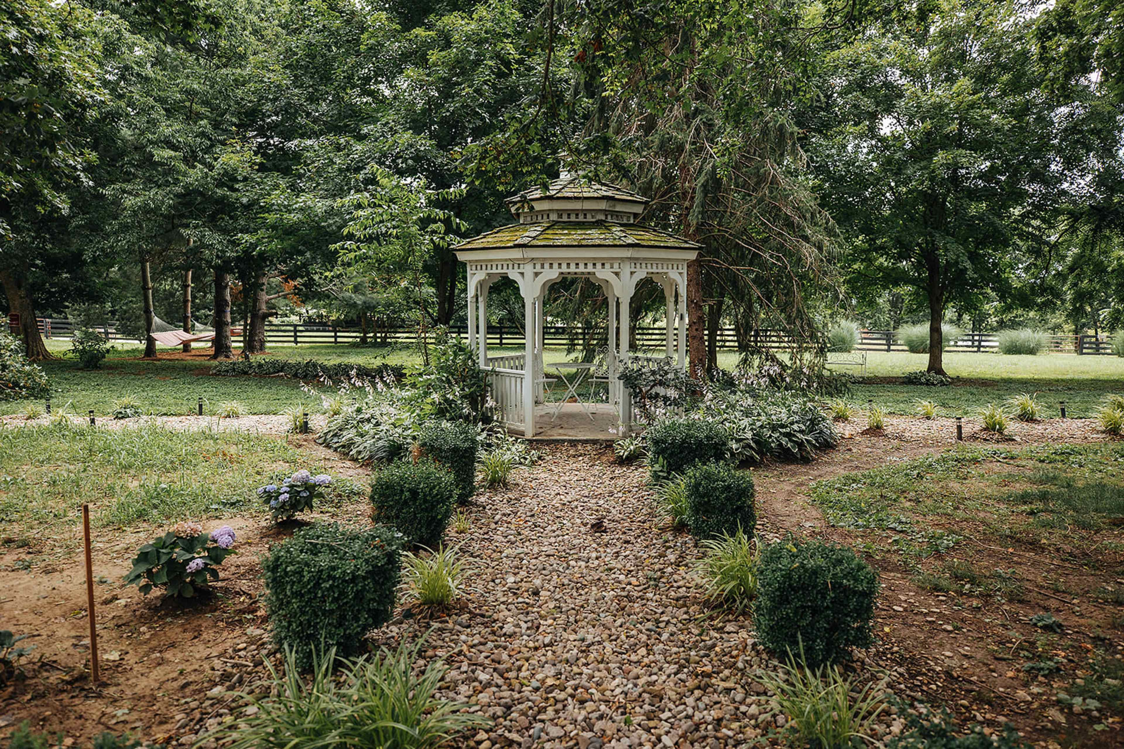 A white gazebo is surrounded by a gravel path and neatly trimmed hedges in a landscaped garden with trees and greenery.
