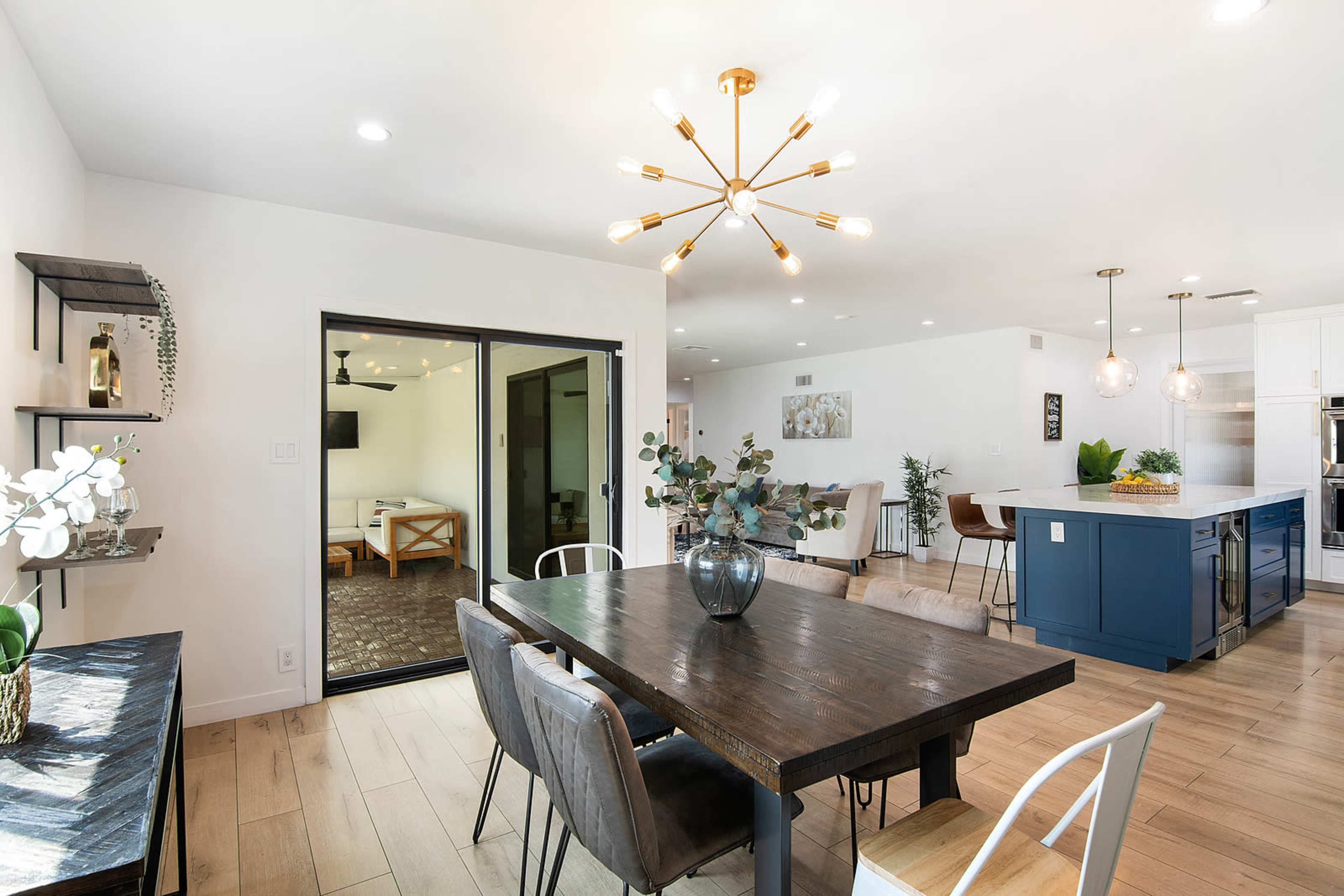 A modern dining area features a wooden table surrounded by chairs, with a sliding glass door leading to an outdoor space and a kitchen island in the background.