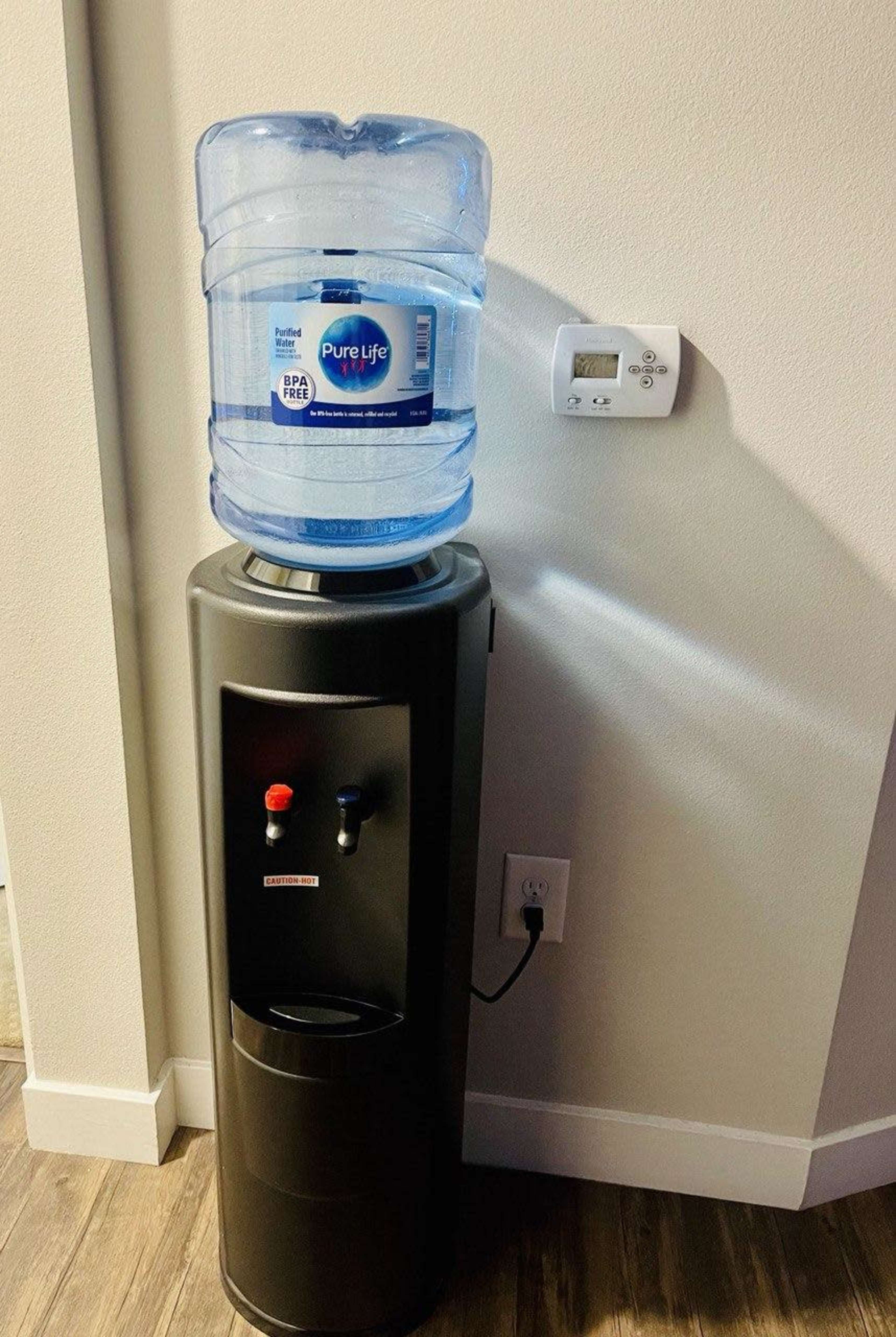 The image shows a water cooler with a large bottle of Pure Life water on top, placed against a wall featuring a thermostat.