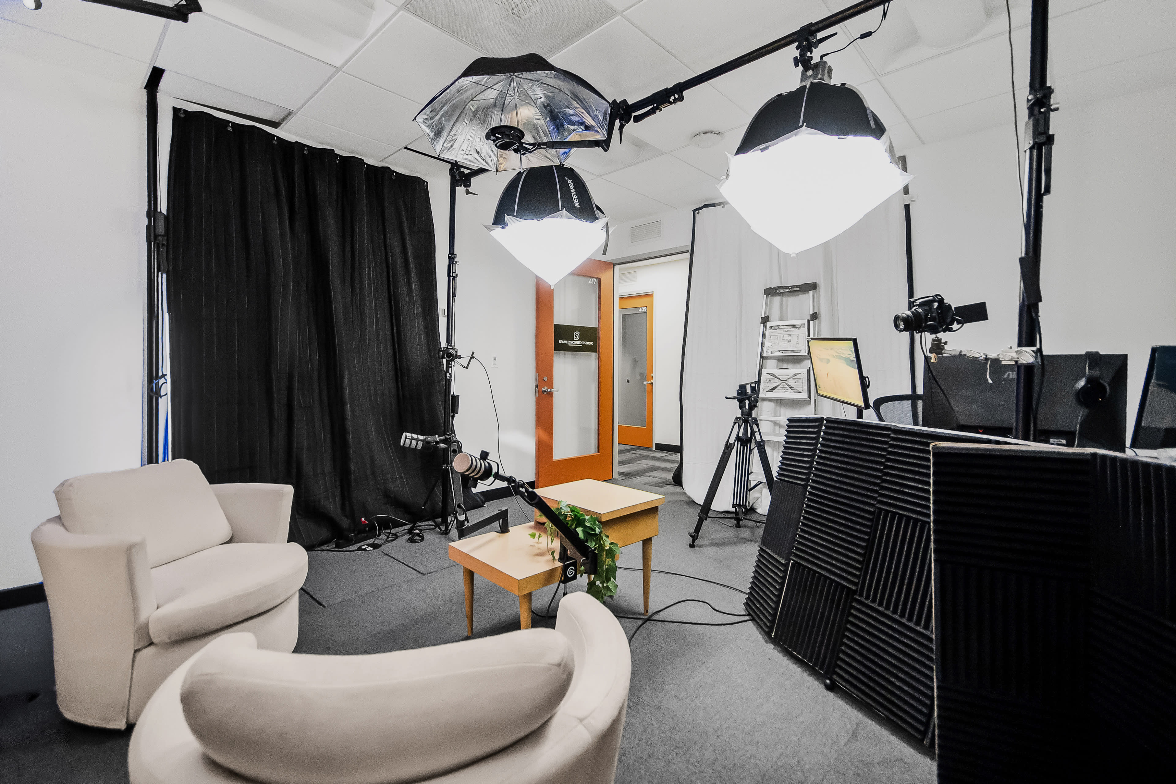 The image shows a small studio setup with two white chairs, a coffee table, and various lighting equipment, along with a backdrop and camera equipment positioned around the space.