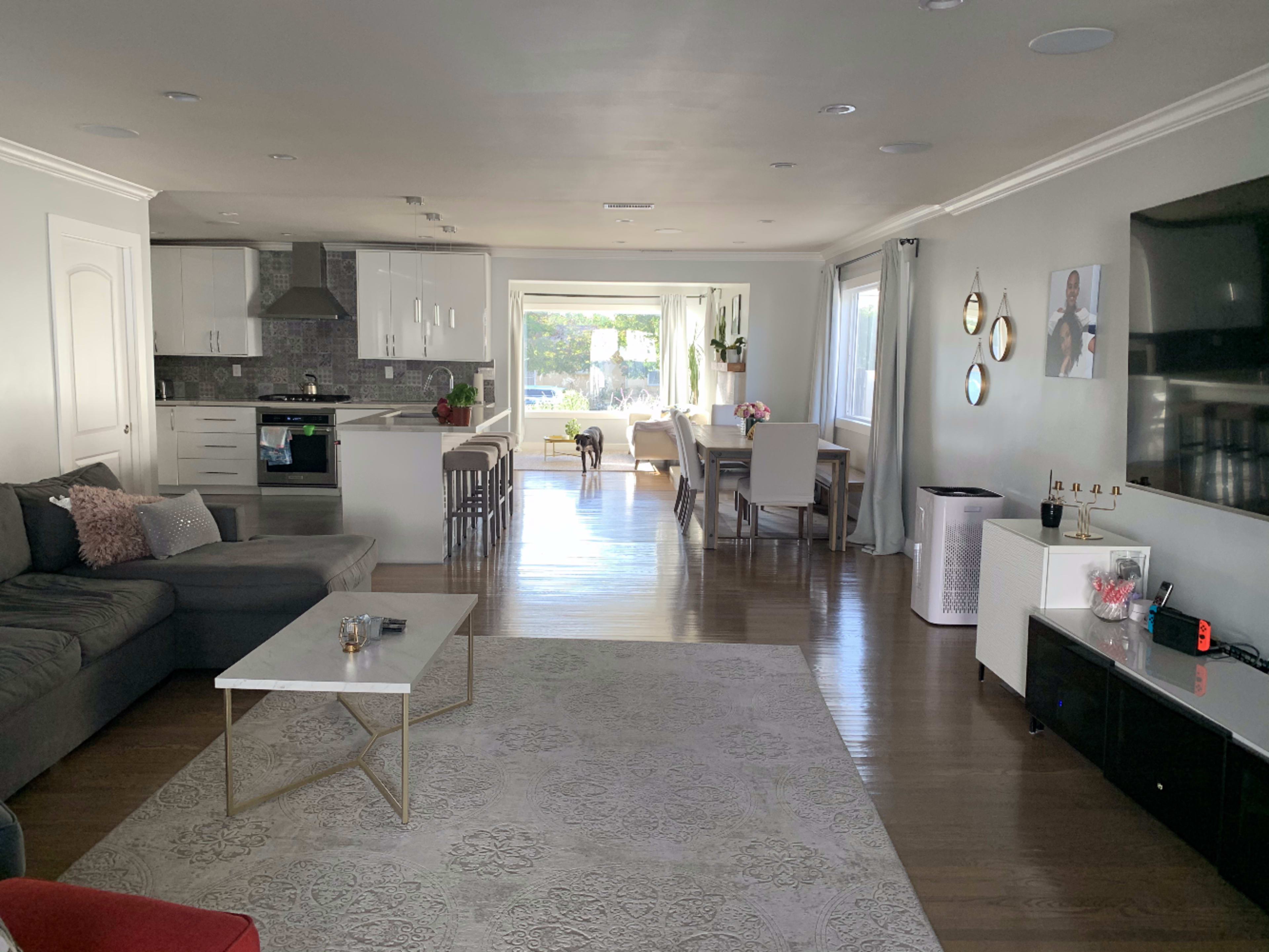 The image shows a modern living room and dining area with a sectional sofa, a coffee table, a kitchen in the background, and large windows letting in natural light.
