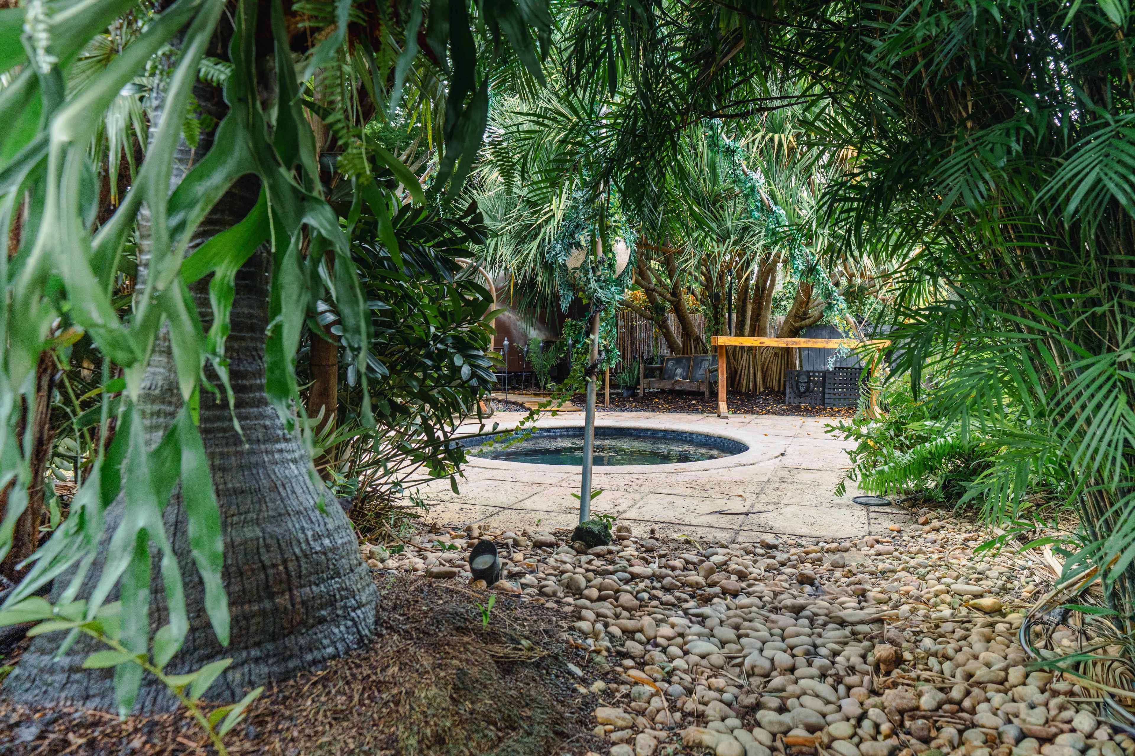 The image shows a secluded backyard with a small, circular pool surrounded by lush greenery and a stone pathway.