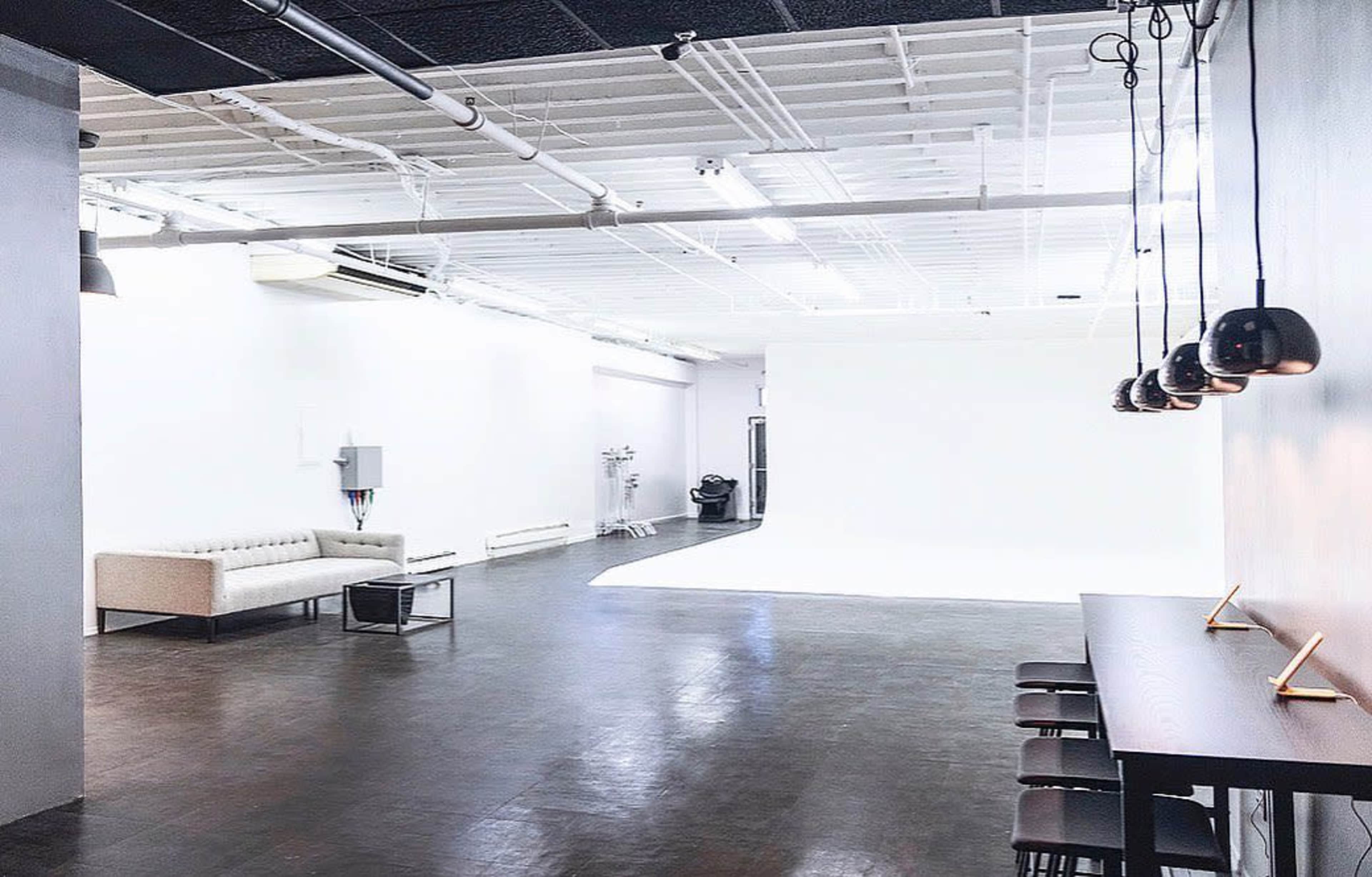 A spacious indoor studio with a white backdrop, a sofa, and a table with chairs, featuring a modern design and industrial lighting.