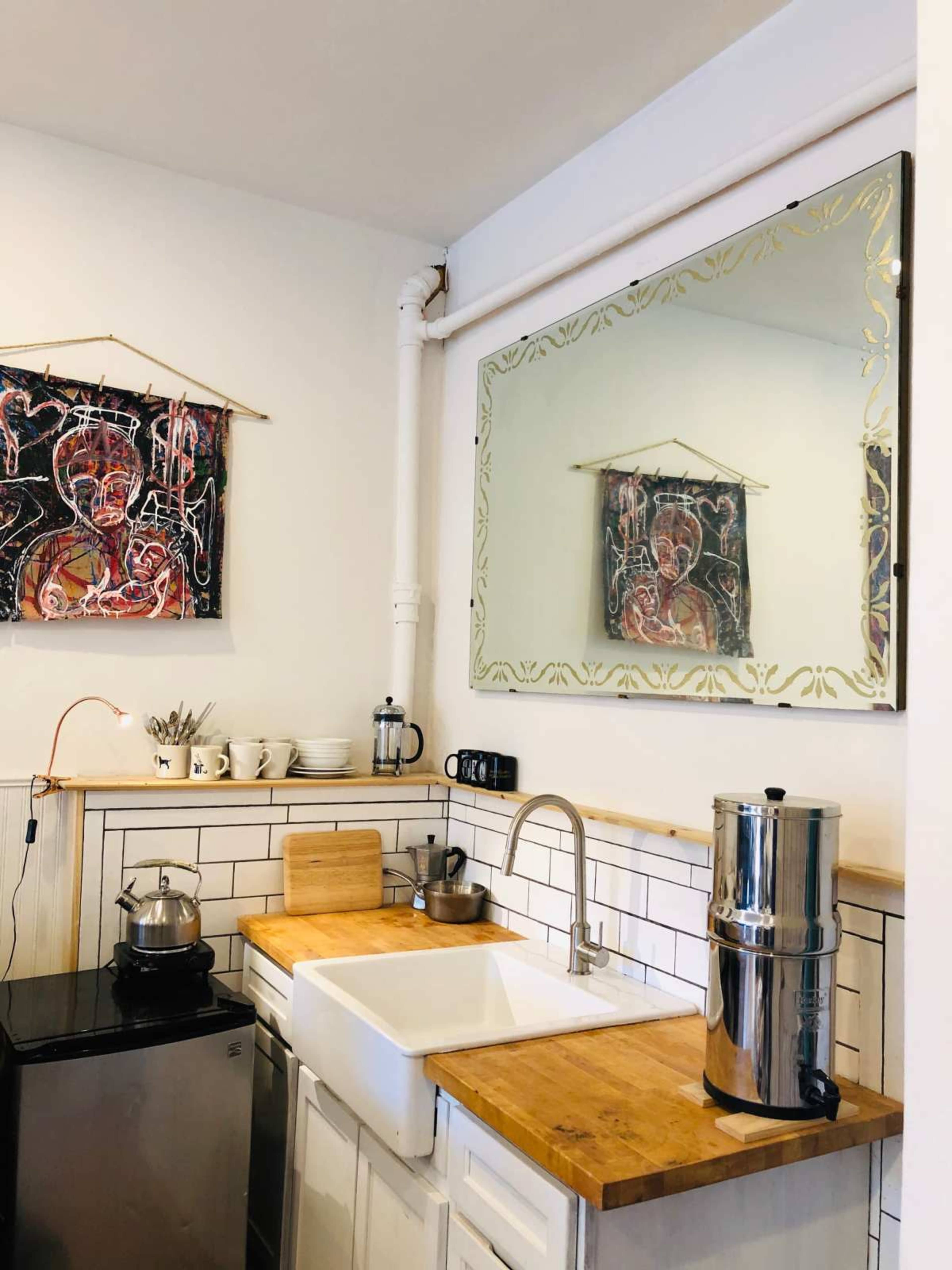 A kitchen features a sink with a wooden countertop, a kettle on the stove, a mirror above, and colorful artwork on the wall.