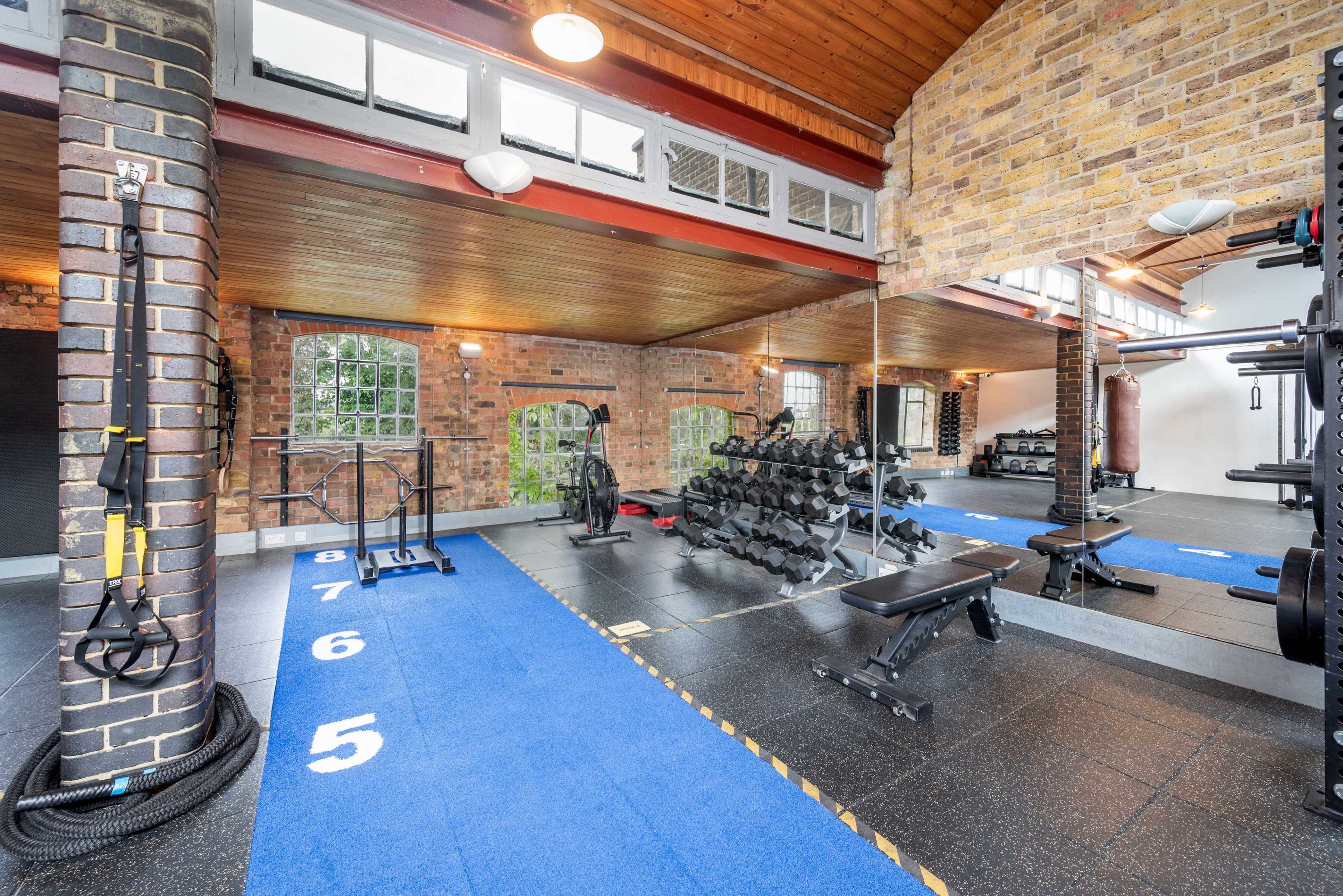 The image shows a modern gym interior with blue carpeting, weightlifting equipment, and large windows letting in natural light.