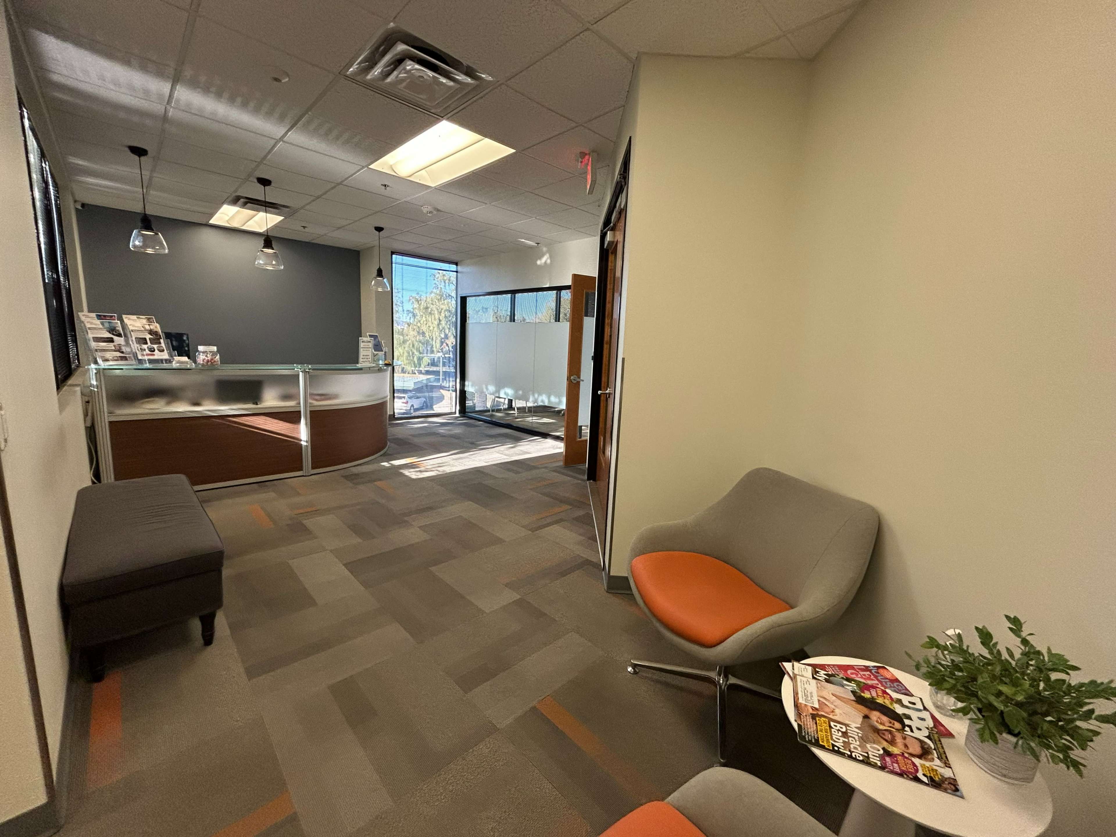 The image shows a modern office interior featuring a reception area with a glass desk, seating arrangements, and large windows providing natural light.