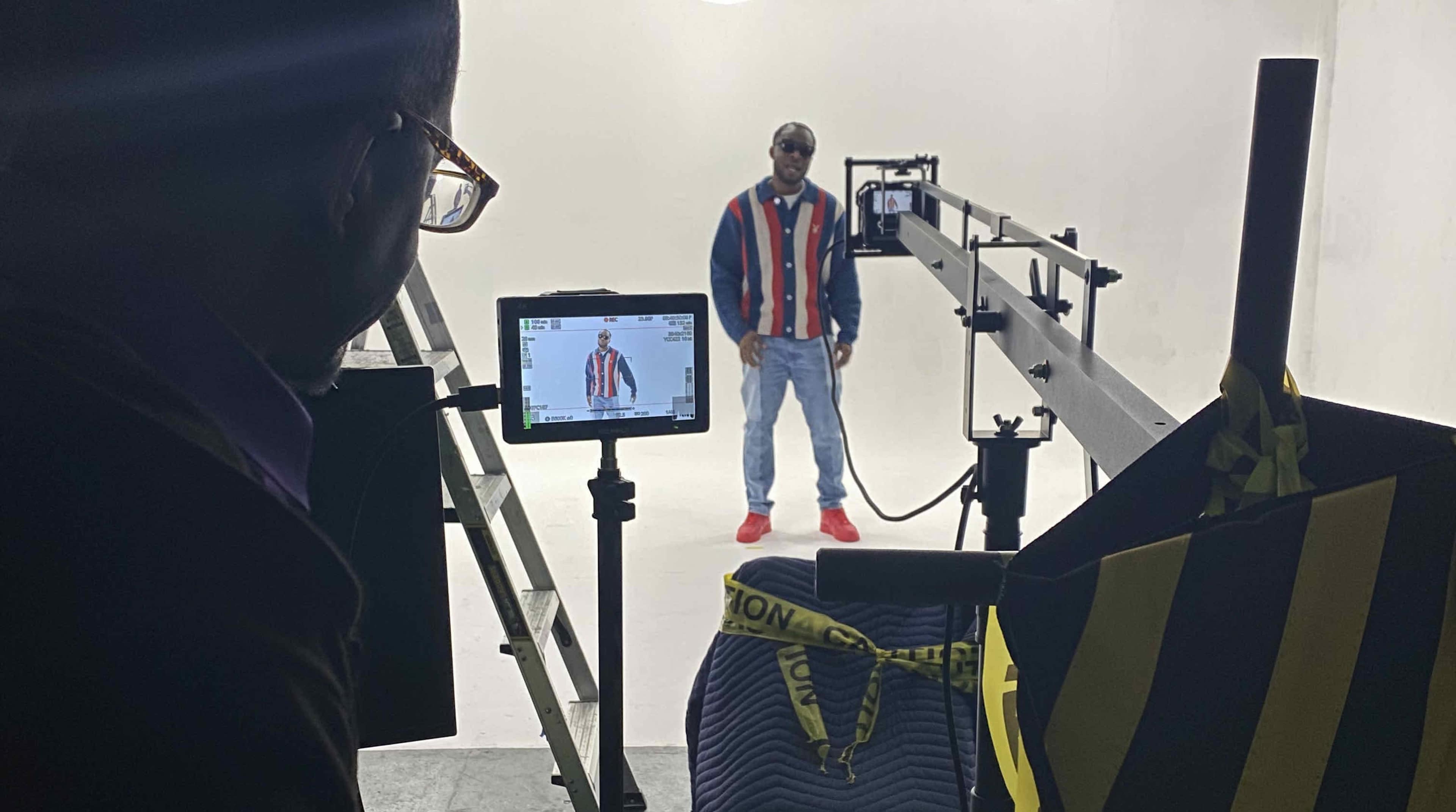 A man dressed in a striped jacket and sunglasses stands in a well-lit studio with filming equipment set up nearby.