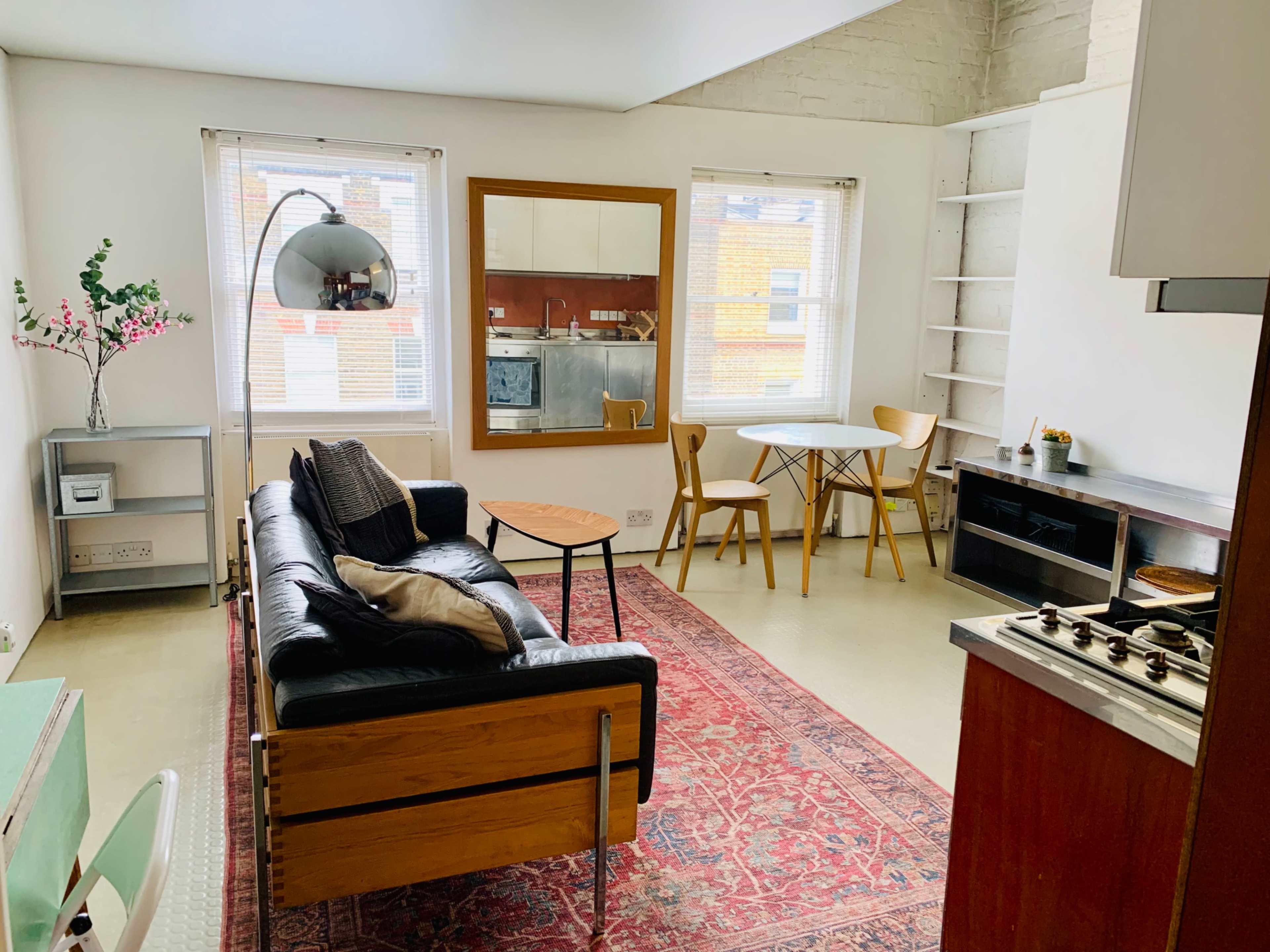 The image shows a modern living room with a black leather sofa, a small wooden table, and a round glass dining table, all set in a bright space with large windows and minimalist decor.
