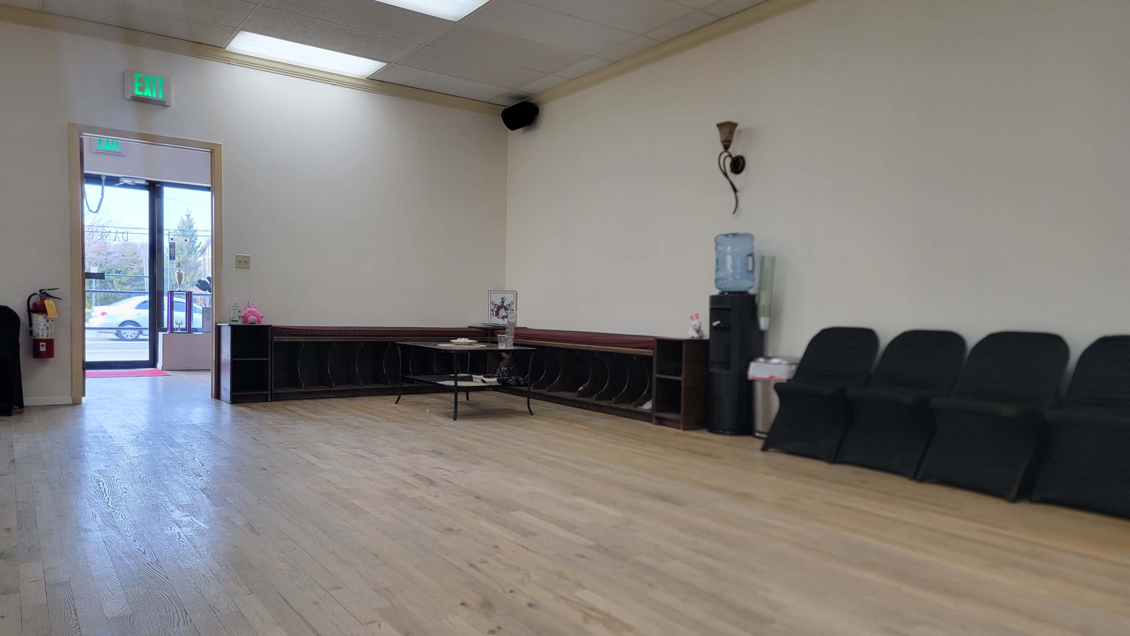 The image shows a spacious, empty room with wooden floors, a water cooler, and a seating area along the walls.