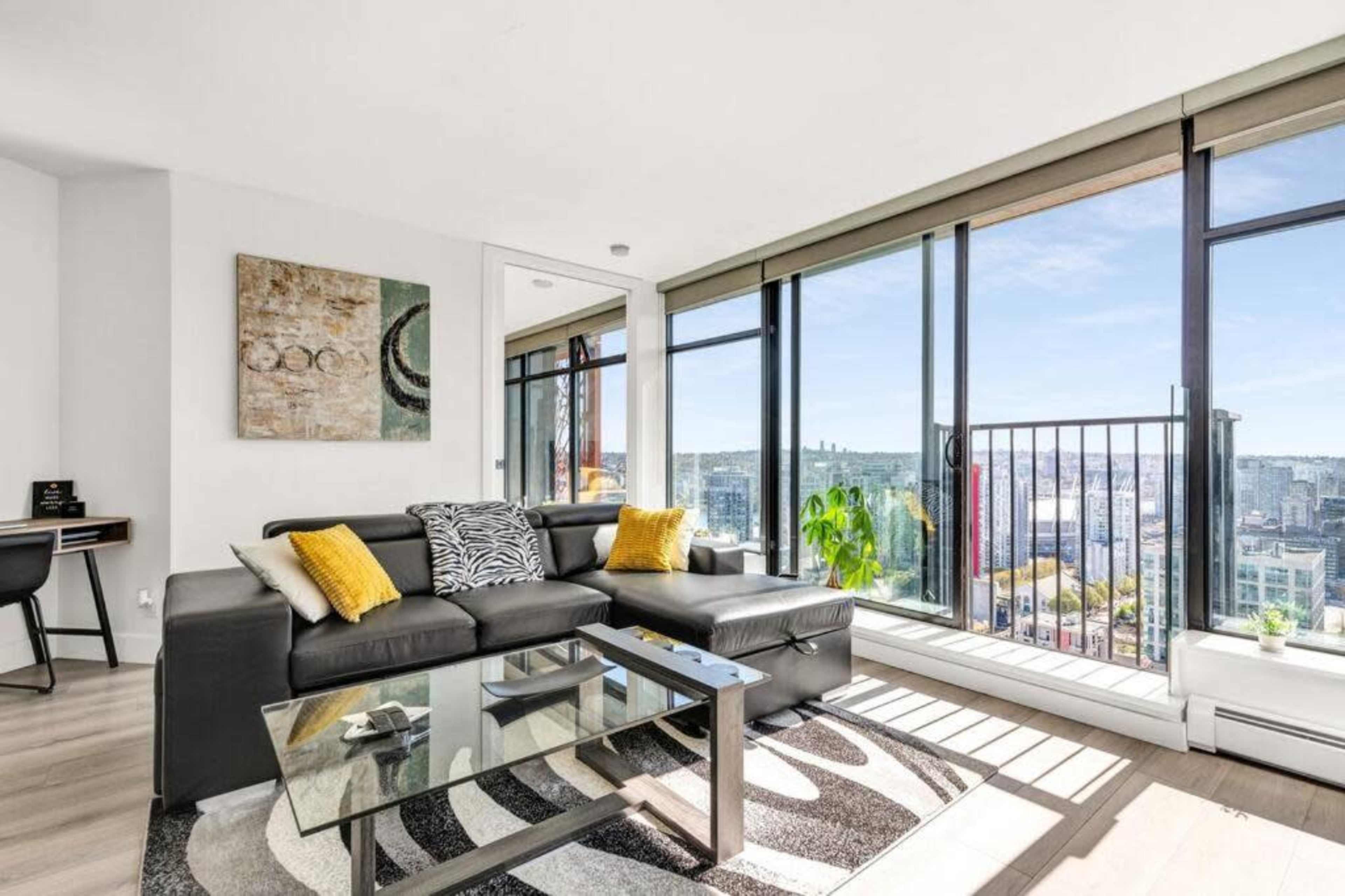 The image shows a modern living room with a black sofa, a glass coffee table, and large windows offering a view of a city skyline.