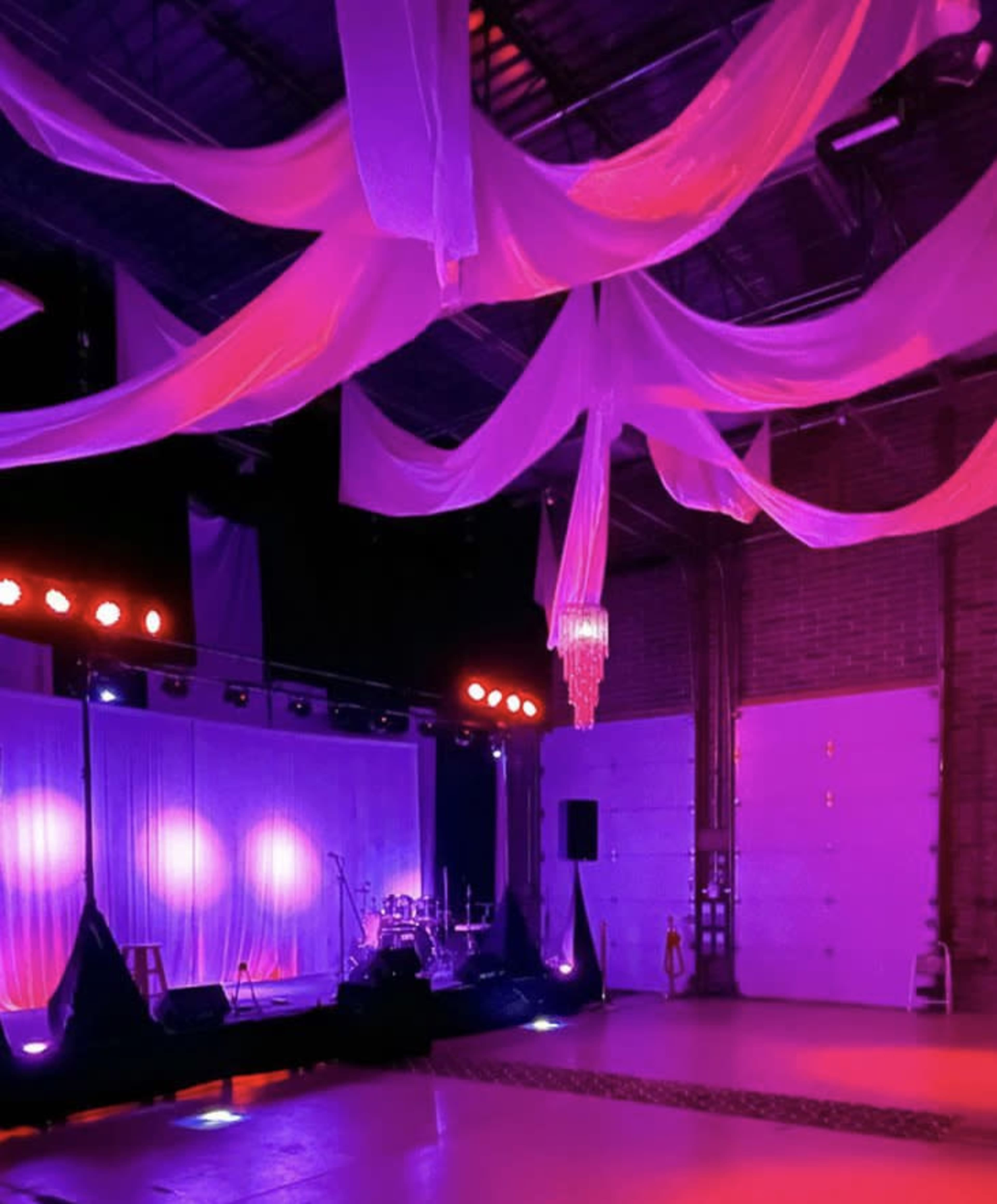 An indoor event space with a stage featuring lighting, draped fabric overhead, and a chandelier.
