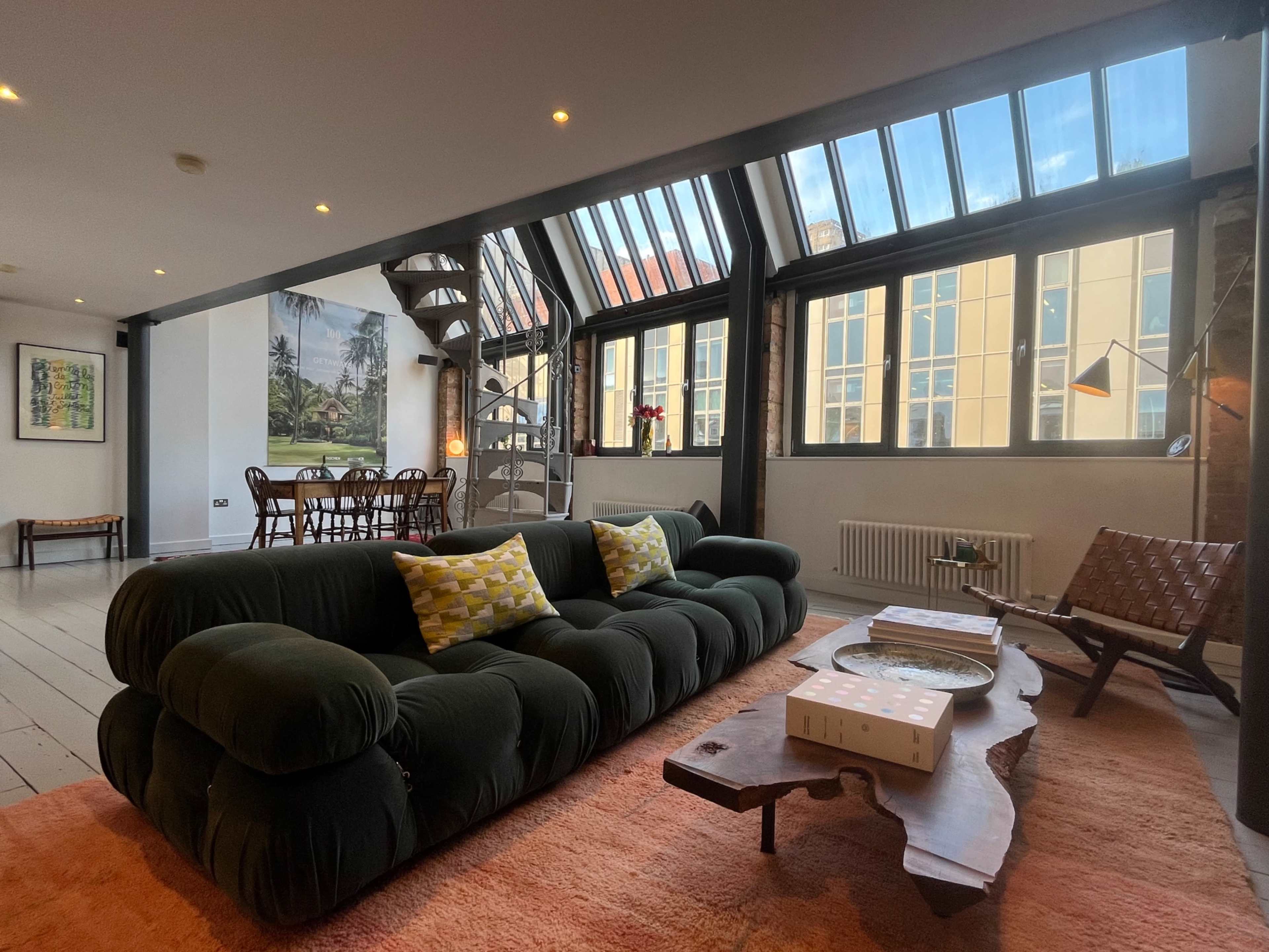 The image shows a modern living room with a large green sofa, a wooden coffee table, and a spiral staircase, all set under high glass ceilings.