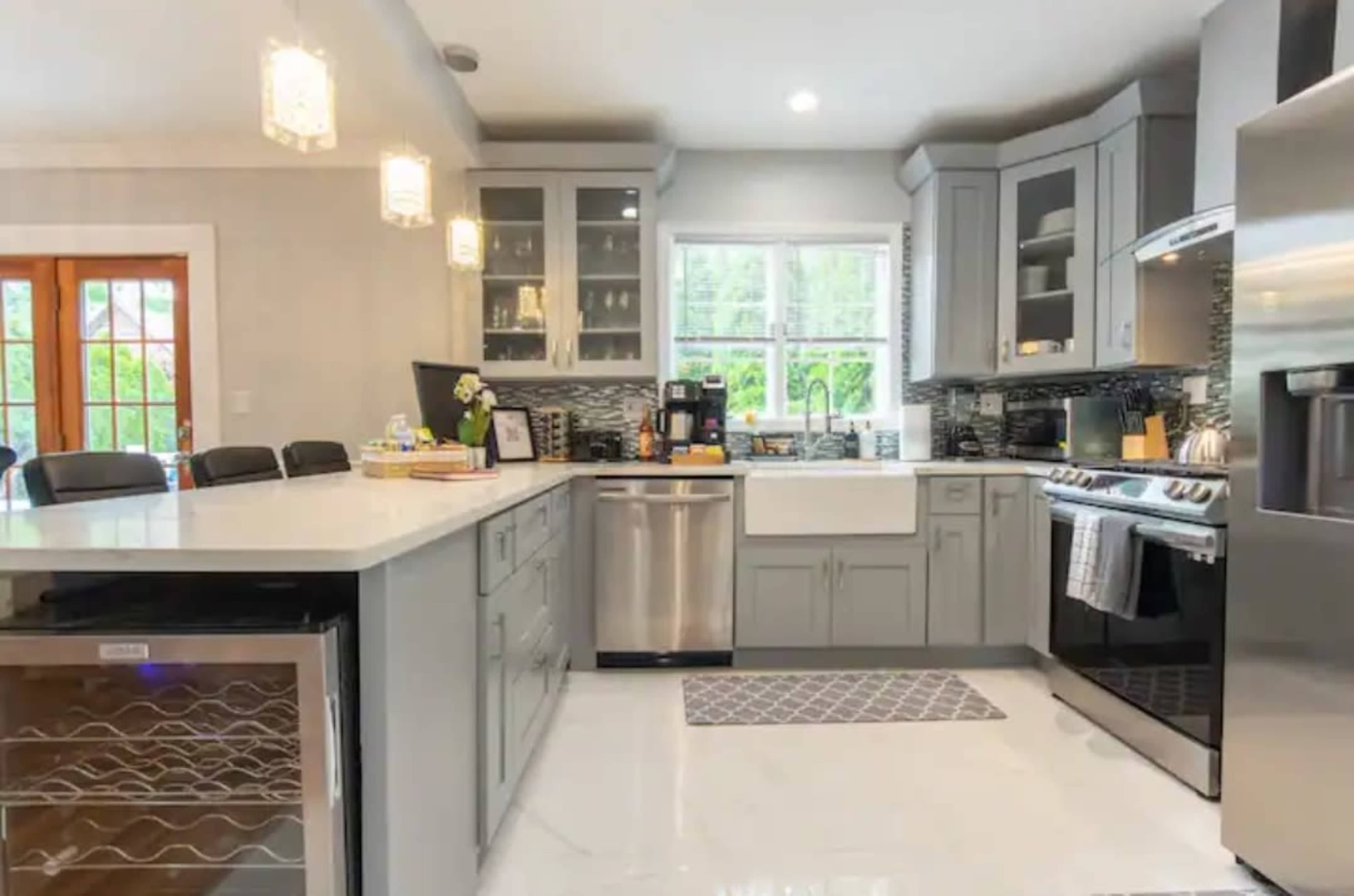 The kitchen features gray cabinets, a stainless steel refrigerator, and a farmhouse sink under a window, with a central island and modern lighting fixtures.