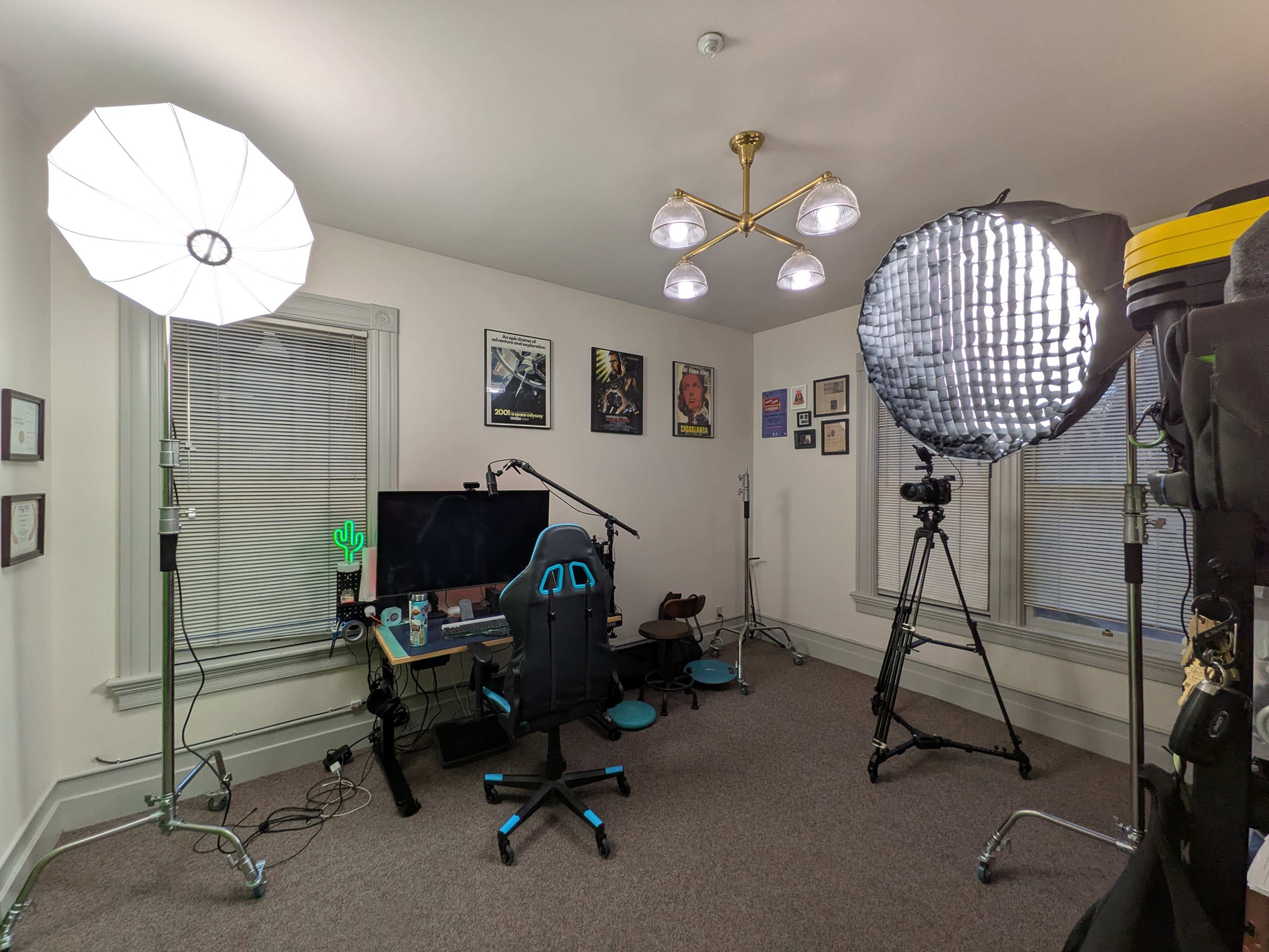 The image shows a well-equipped home studio featuring a black gaming chair, a large monitor, two softbox lights, and a tripod, all set up in a room with light-colored walls and windows.