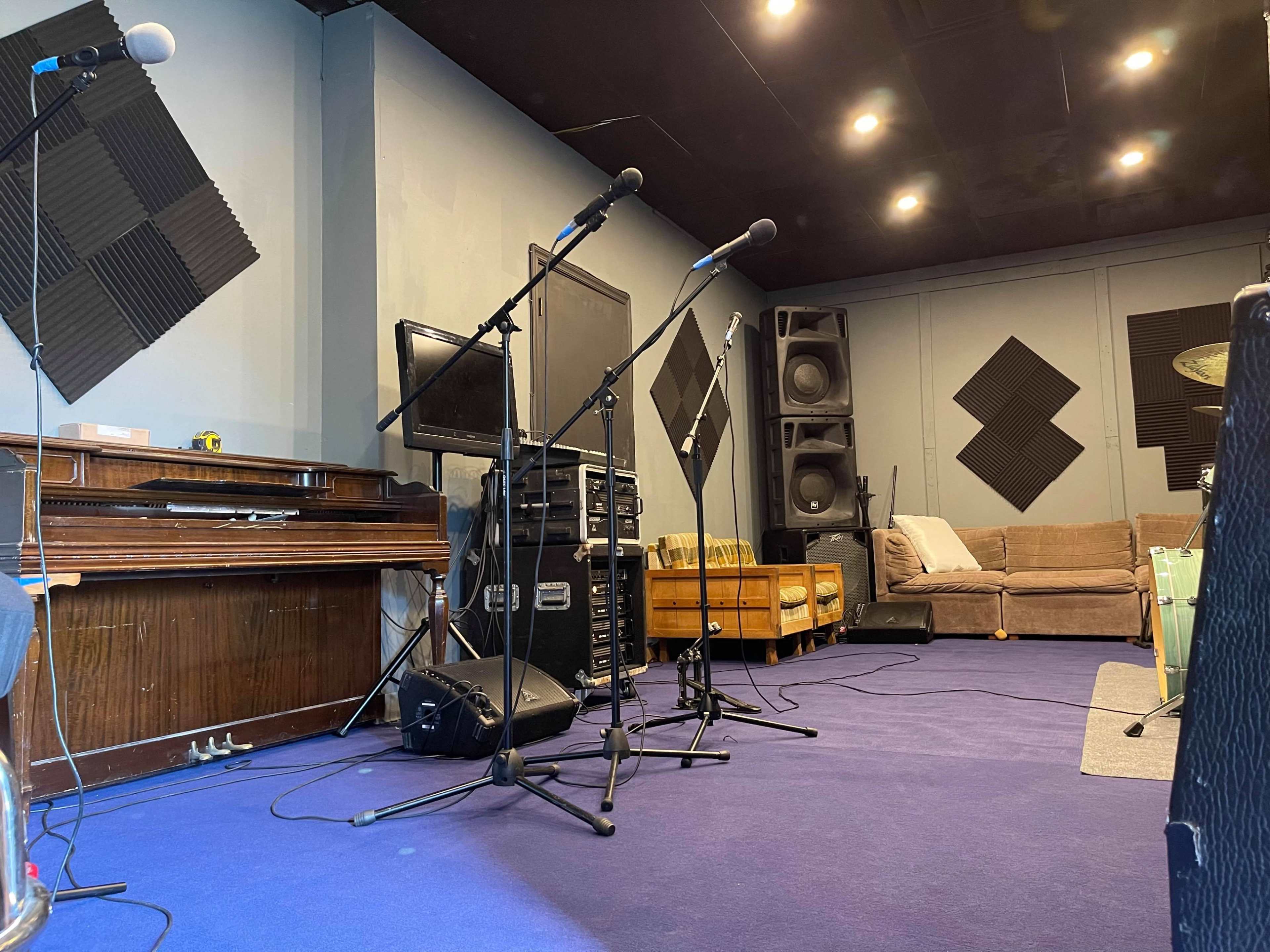 A music studio with microphones set up in front of a piano, a stack of sound equipment, and a couch in the background.