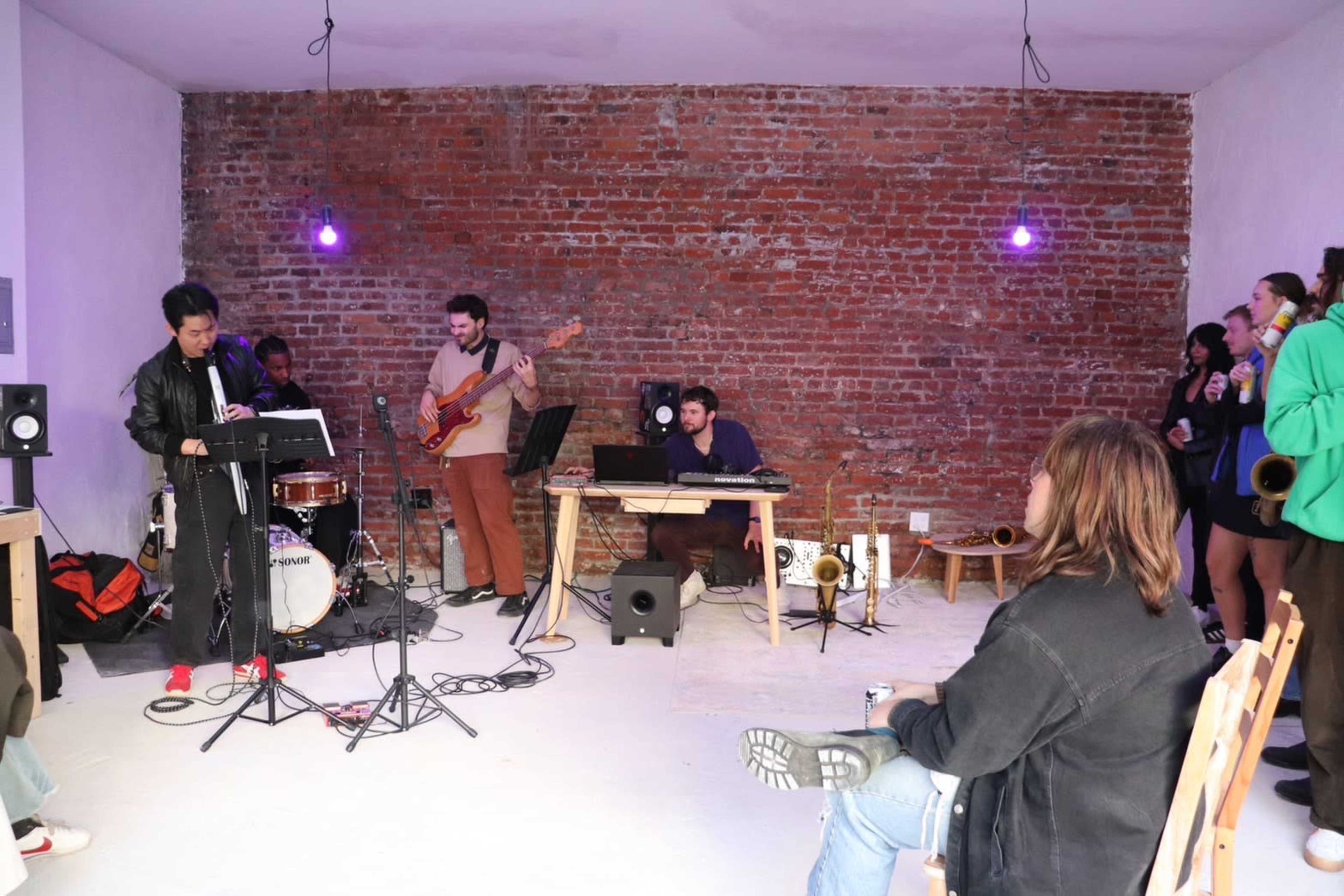 A band performs in a room with exposed brick walls, featuring various musicians playing instruments and a small audience gathered around.
